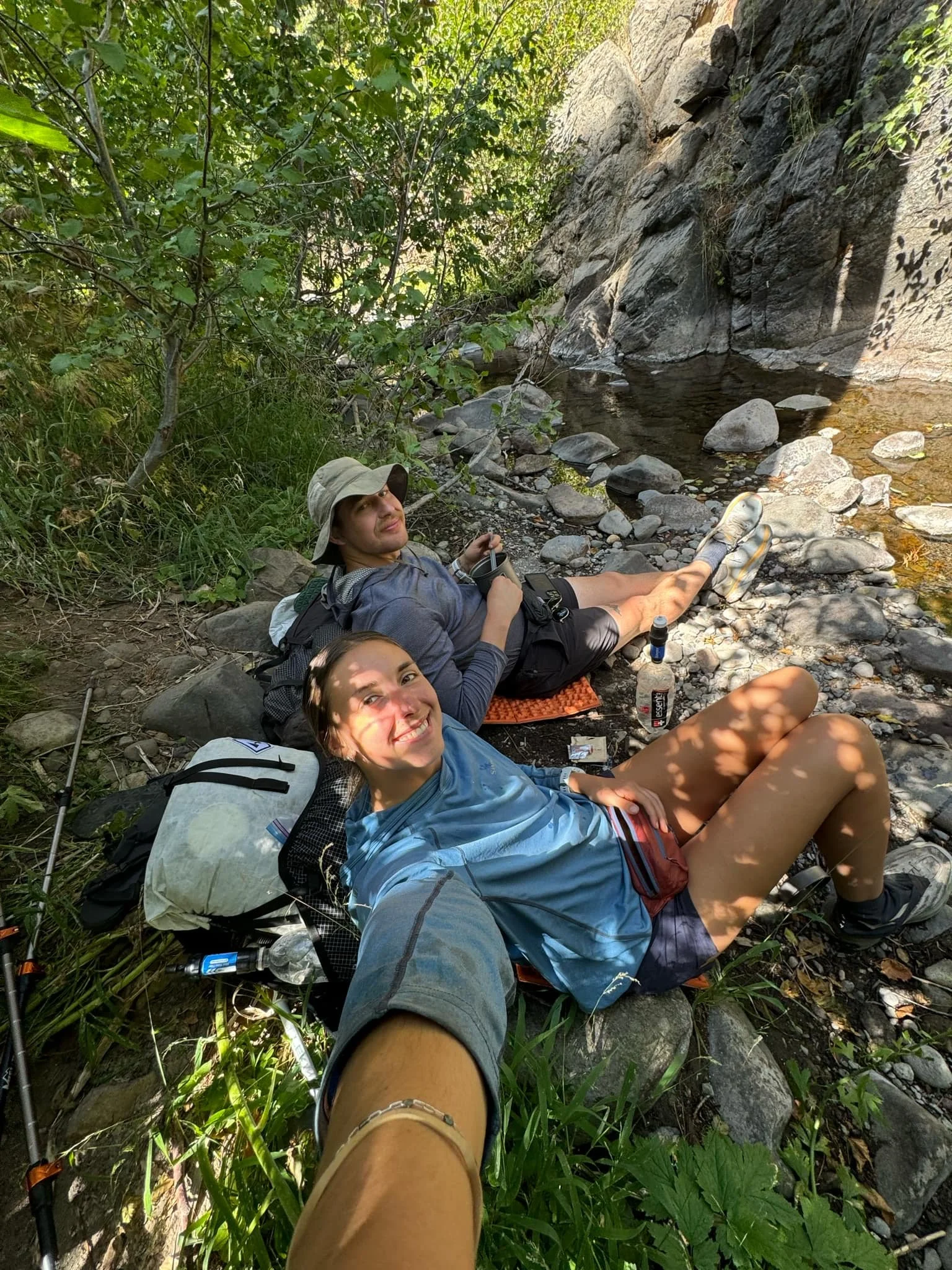 Two people relaxing by a rocky creek in a wooded area, lying on the ground with backpacks and gear around them.