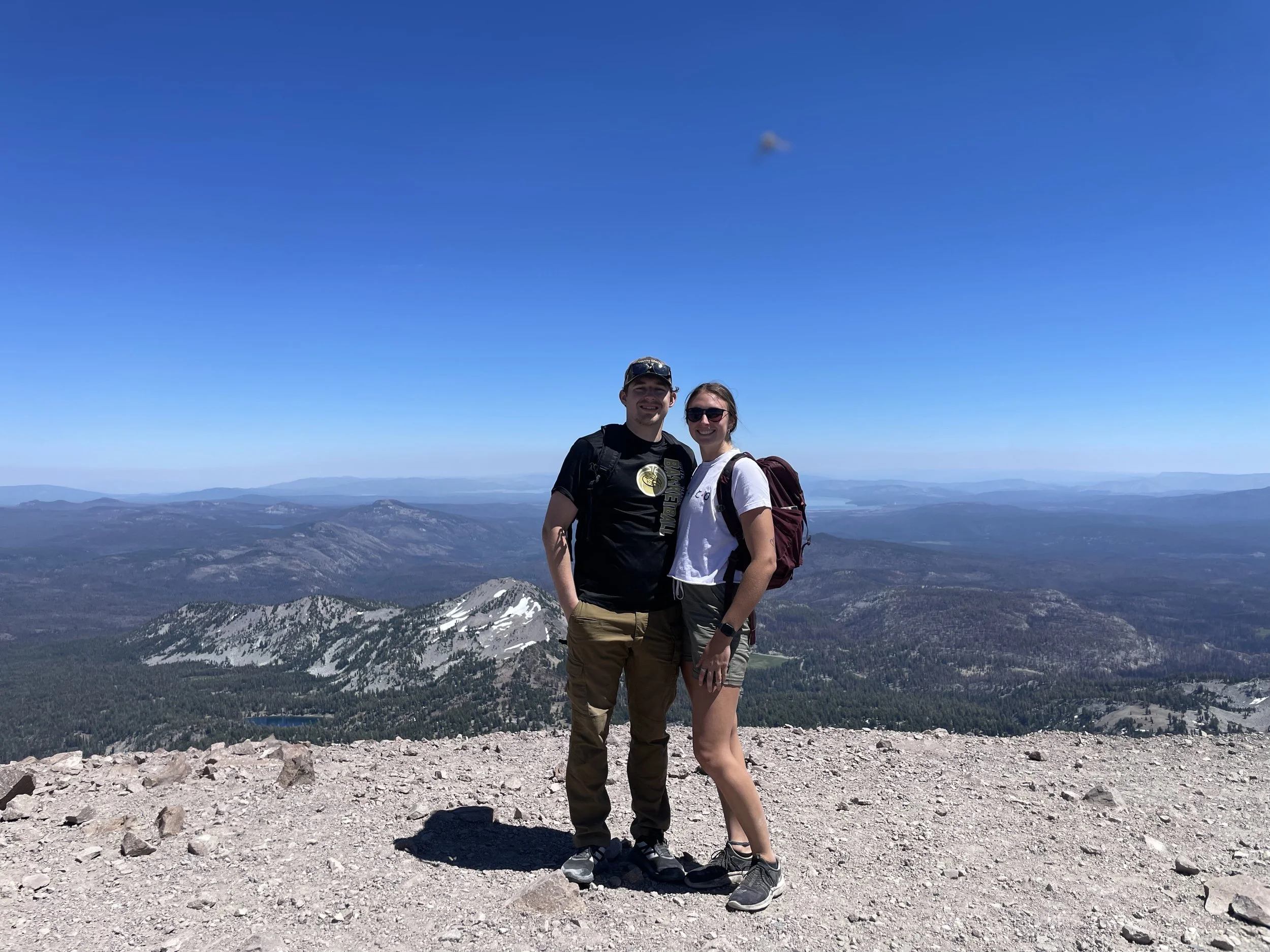 A man and woman standing together on a mountain summit with a panoramic view of mountains and valleys in the background, bright blue sky above.