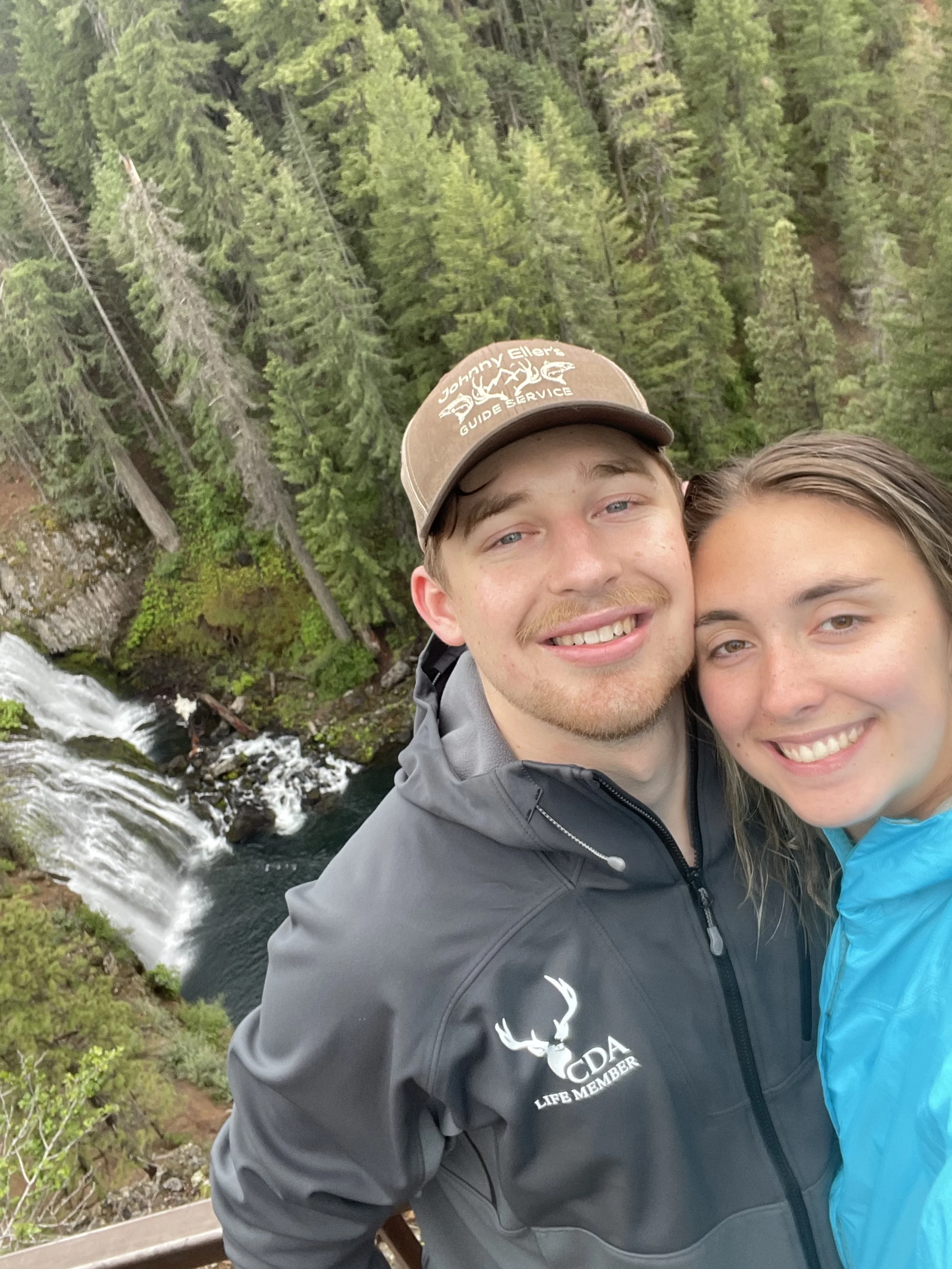 A smiling young couple taking a selfie outdoors near a forest with tall green trees and a waterfall in the background.