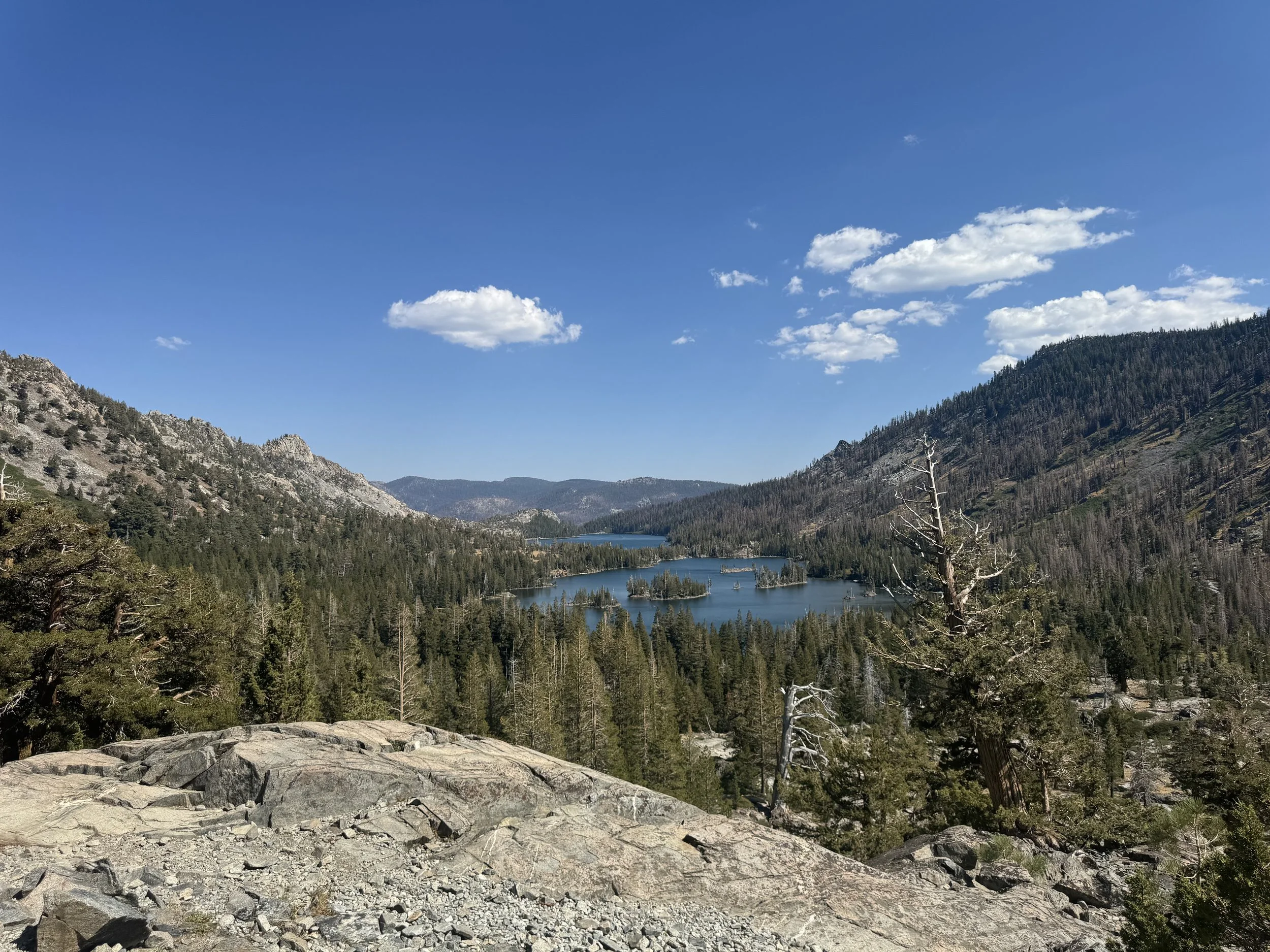 A scenic view of a mountain lake surrounded by dense pine forests and rocky mountain slopes under a blue sky with scattered clouds.
