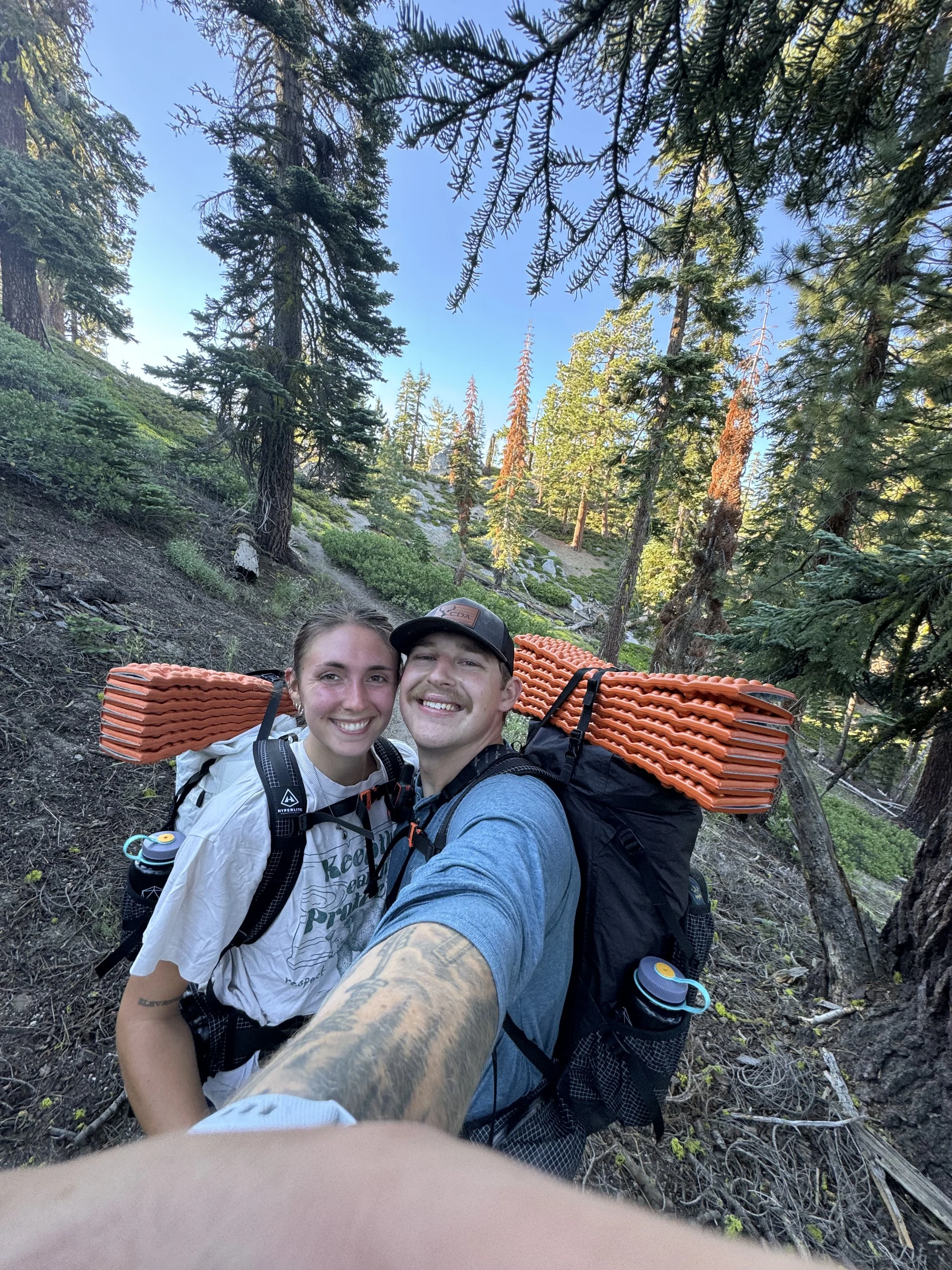 A smiling couple taking a selfie during a hike in a forested area with tall pine trees and a clear blue sky.