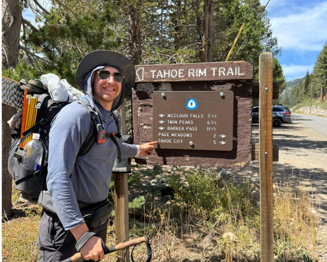 A man in outdoor hiking gear, including sunglasses, a wide-brim hat, and a backpack with supplies, points at a trail sign for the Tahoe Rim Trail.