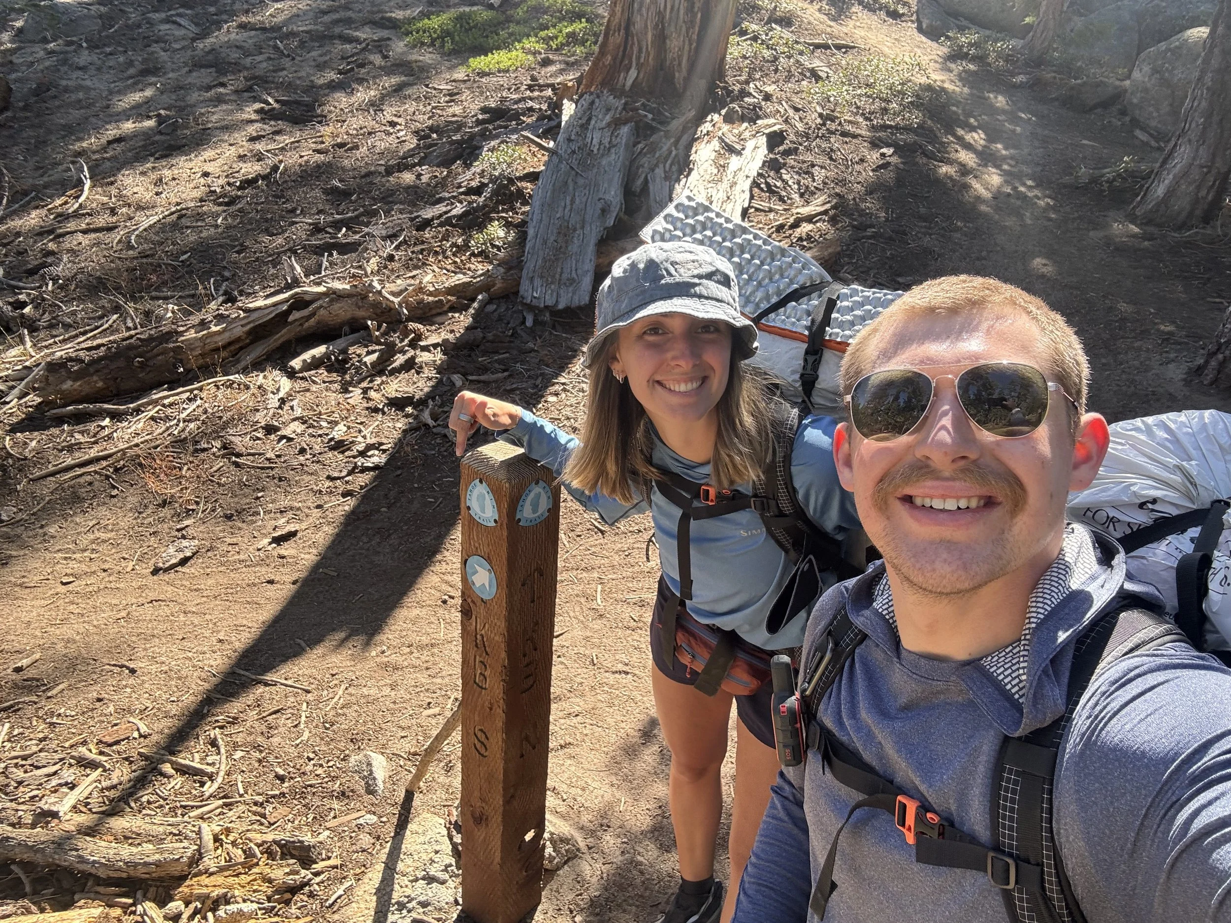 A young man and woman hiking on a dirt trail in the woods, smiling for a selfie. The woman is wearing a hat and sunglasses, while the man is wearing sunglasses. They are carrying backpacks, and there is a trail marker post with directional signs besi
