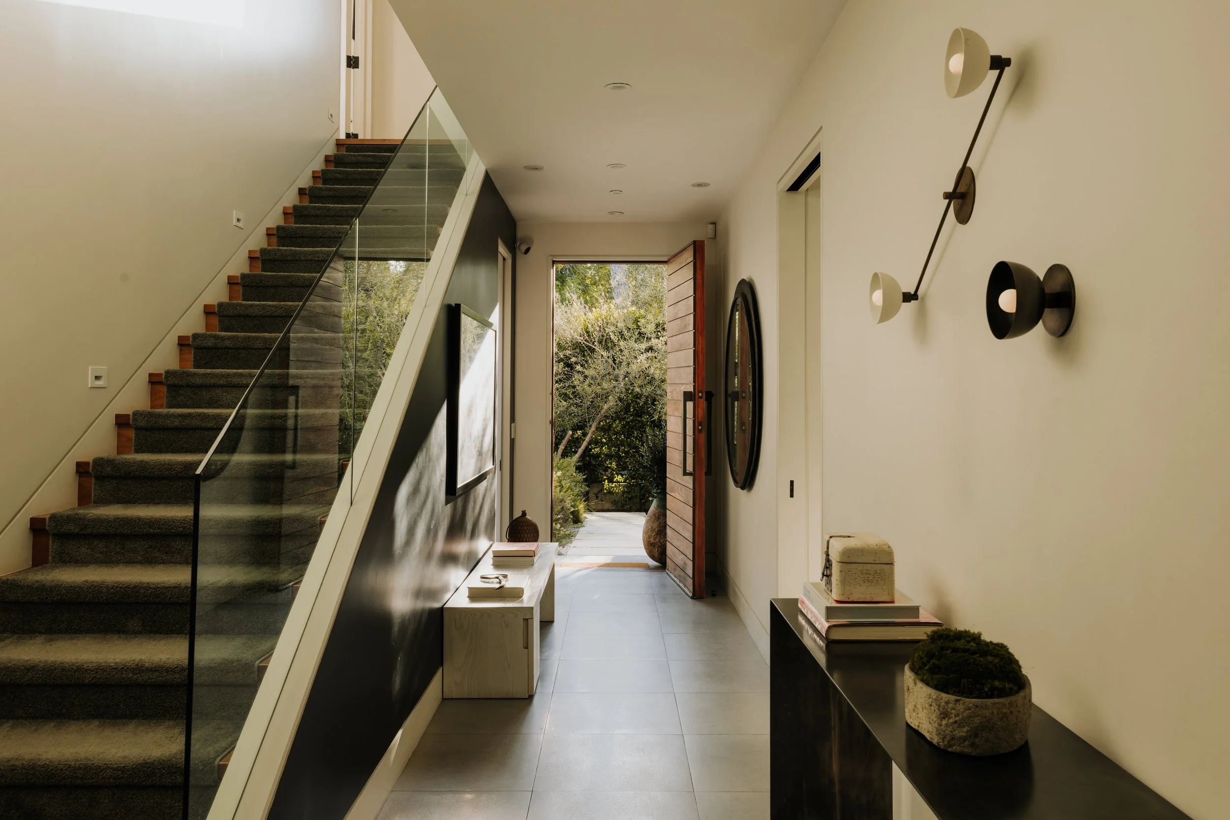 Entrance hall with carpeted stairs, open wooden door, view of the outdoor garden, and modern décor featuring shelves and decorative plants.