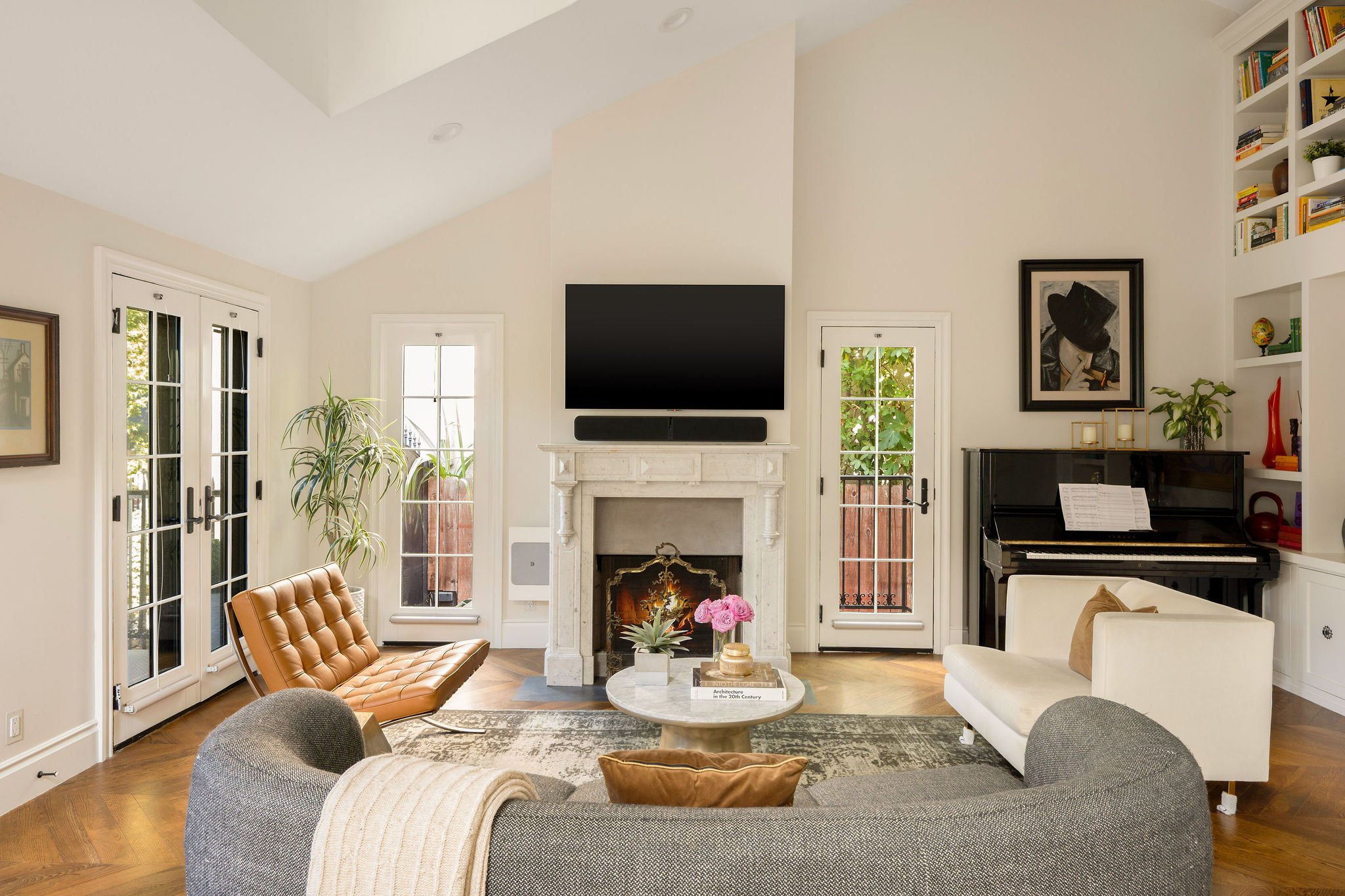Living room with a stone fireplace, flat-screen TV, wicker armchair, gray sofa, desk with piano, white bookshelf with books and decorations, glass doors and windows, plant, wall art, and a rug over a wooden floor.