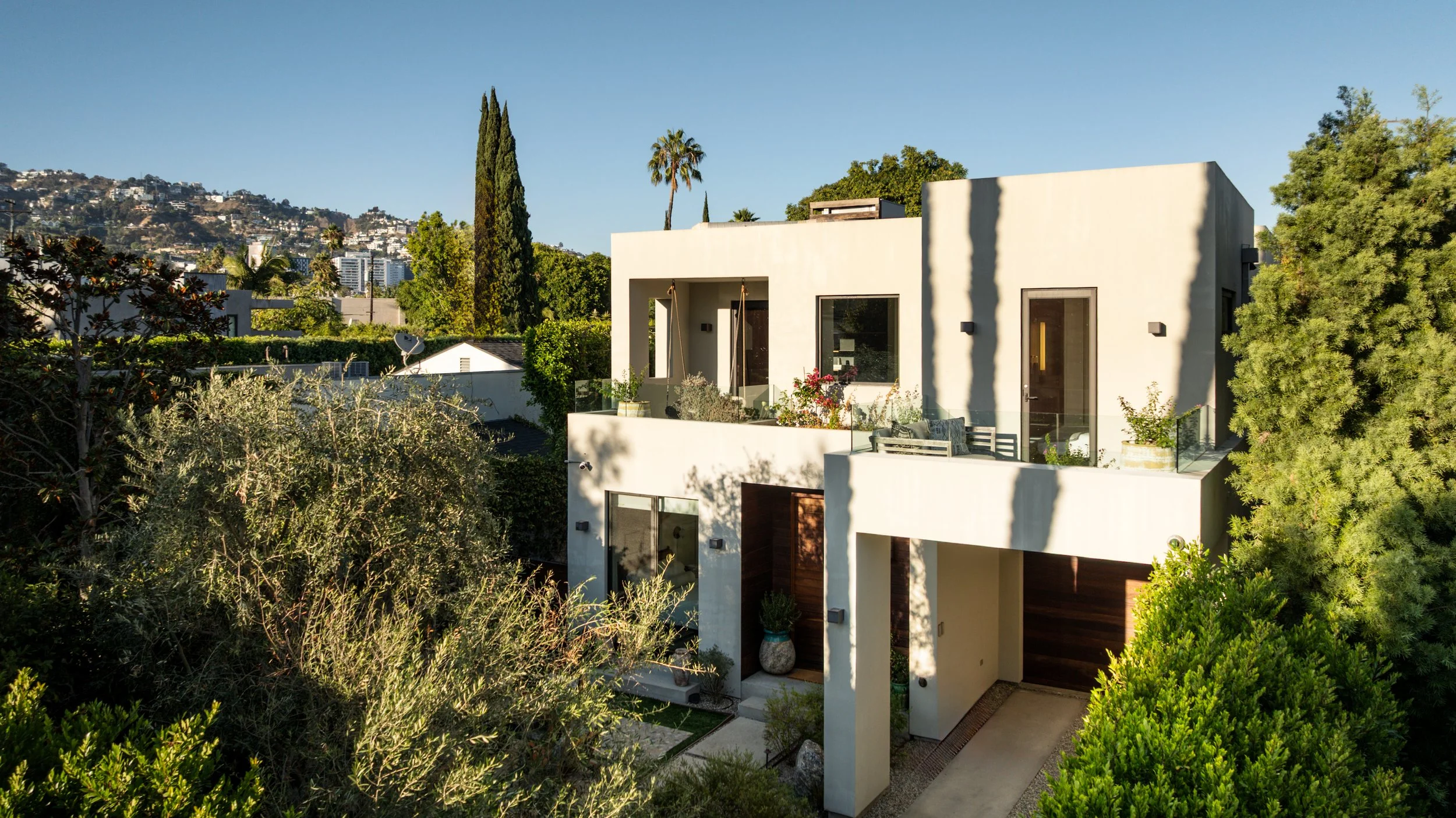 Modern two-story house with balconies and gardens, surrounded by trees and greenery on a sunny afternoon.