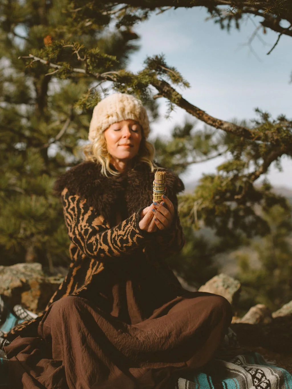 A woman sitting outdoors among trees, holding a bundle of sage, wearing a knit hat, animal print coat, and brown pants, with a peaceful expression on her face.
