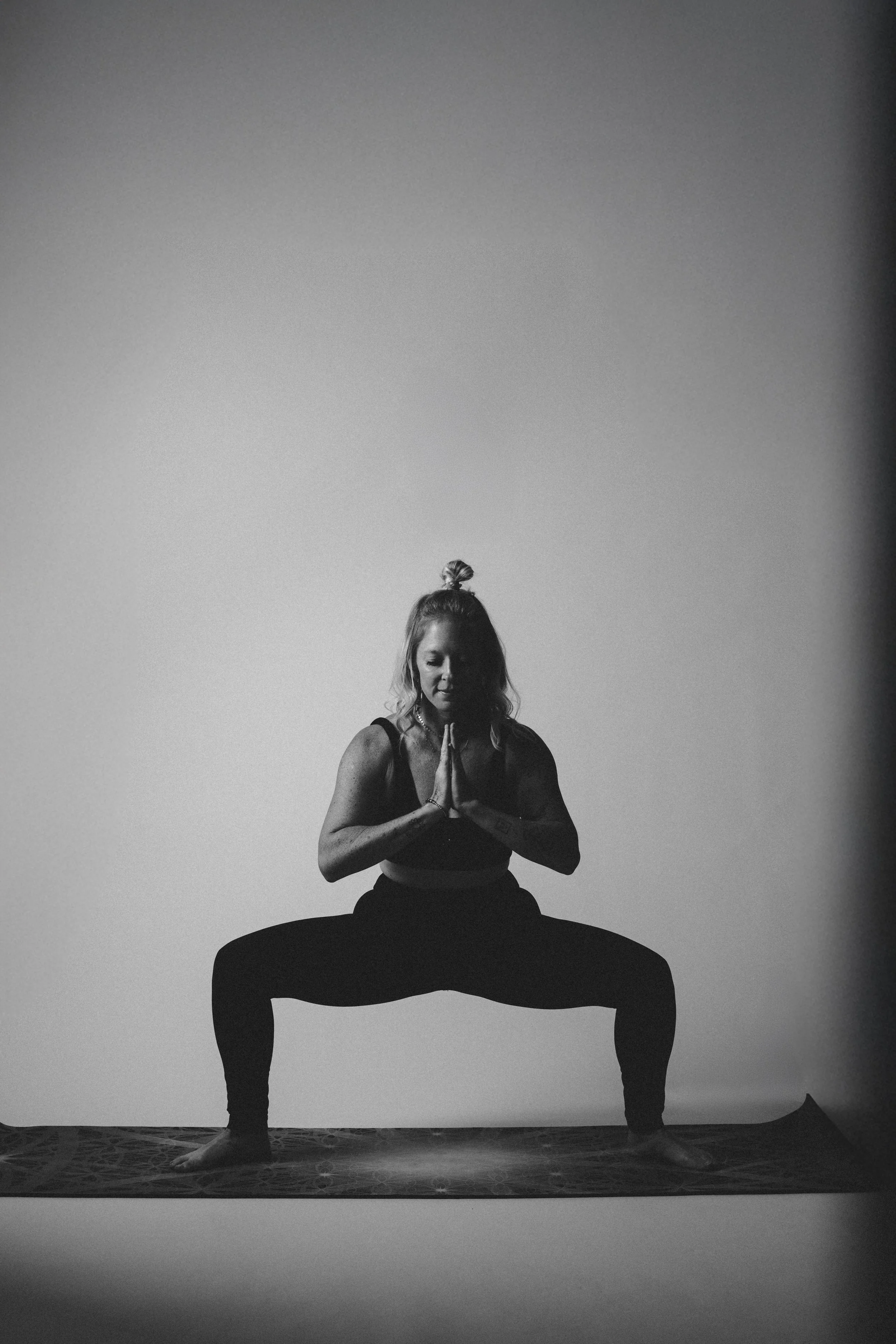 A woman practicing yoga in a squat pose on a yoga mat, with her hands in prayer position, black and white photo.