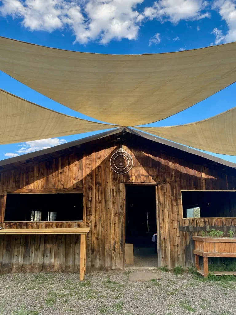 Wooden barn with open door and windows, shaded by two large beige fabric canopies against a blue sky with scattered clouds.