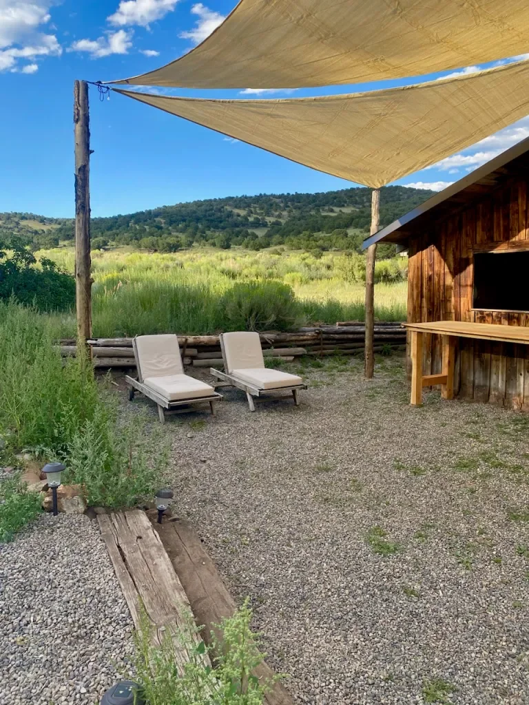 Two lounge chairs with beige cushions facing a scenic green landscape under a large beige shade sail supported by wooden poles, next to a wooden structure with outdoor lighting and a small wooden counter, set on a gravel ground with a wooden plank walkway.