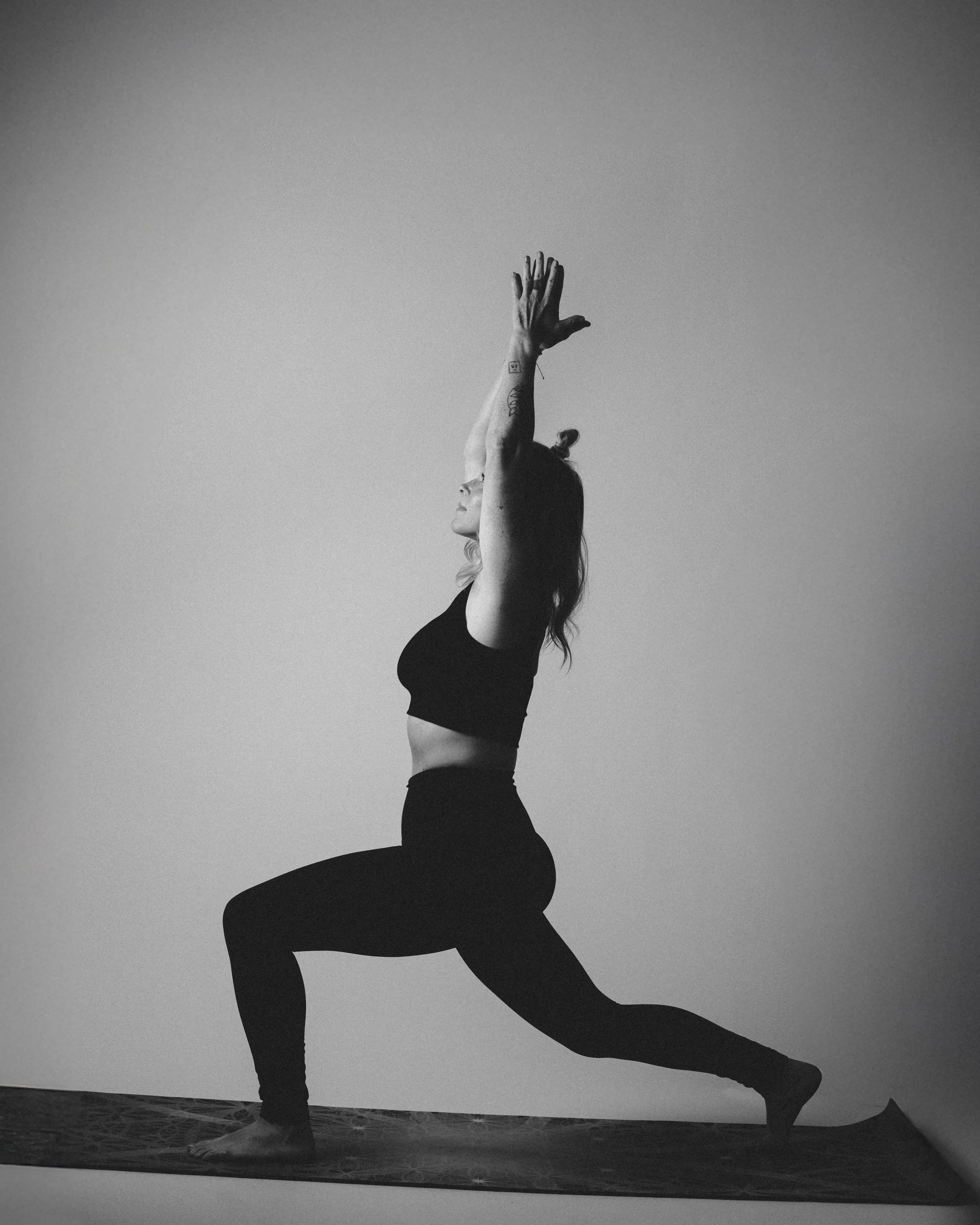 Black and white photo of a woman in yoga pose on a mat, with one leg forward and bent, the other extended back, arms raised above her head, facing the side.