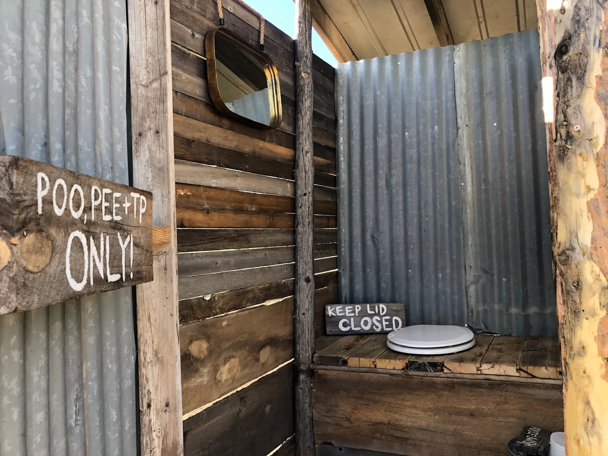 An outdoor rustic outhouse with corrugated metal and wooden walls, a mirror, and a toilet with signs reading 'Poop Pee + TP only' and 'Keep lid closed'.