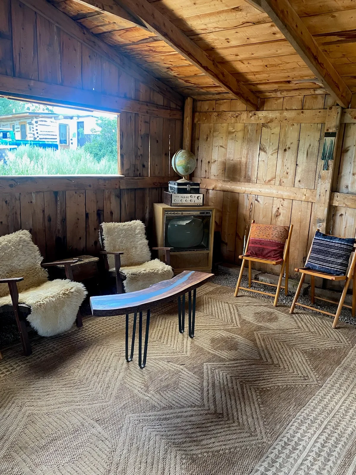 Cozy rustic room with wooden walls and ceiling, featuring a window showing greenery outside, vintage TV and stereo, globe, and chairs with furry and fabric cushions, along with a wooden coffee table and patterned rug.