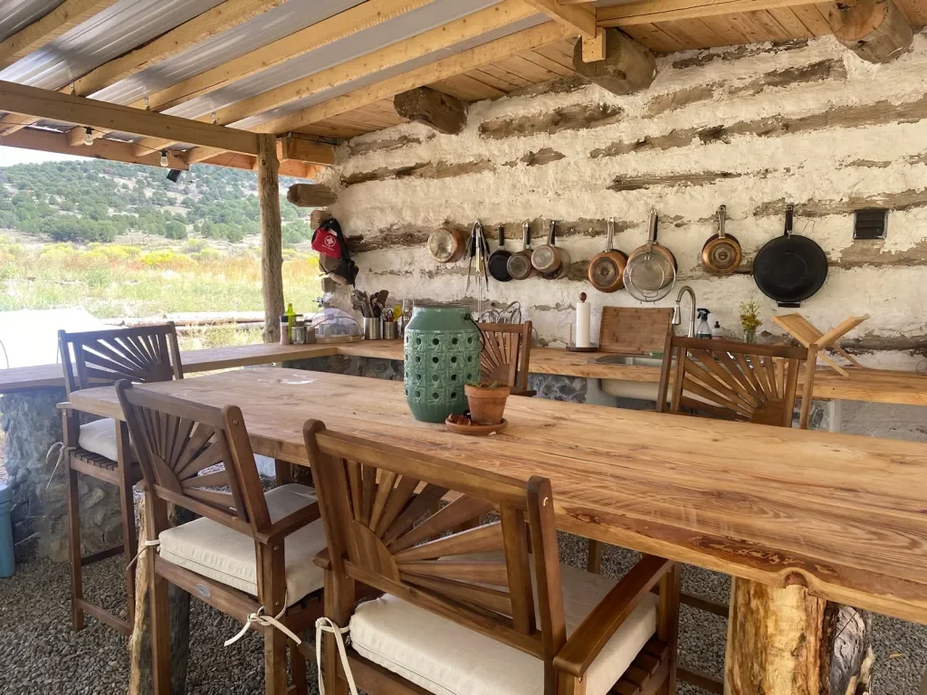 Rustic outdoor kitchen with a long wooden table, matching wooden chairs, and a stone wall with hanging pots and pans.