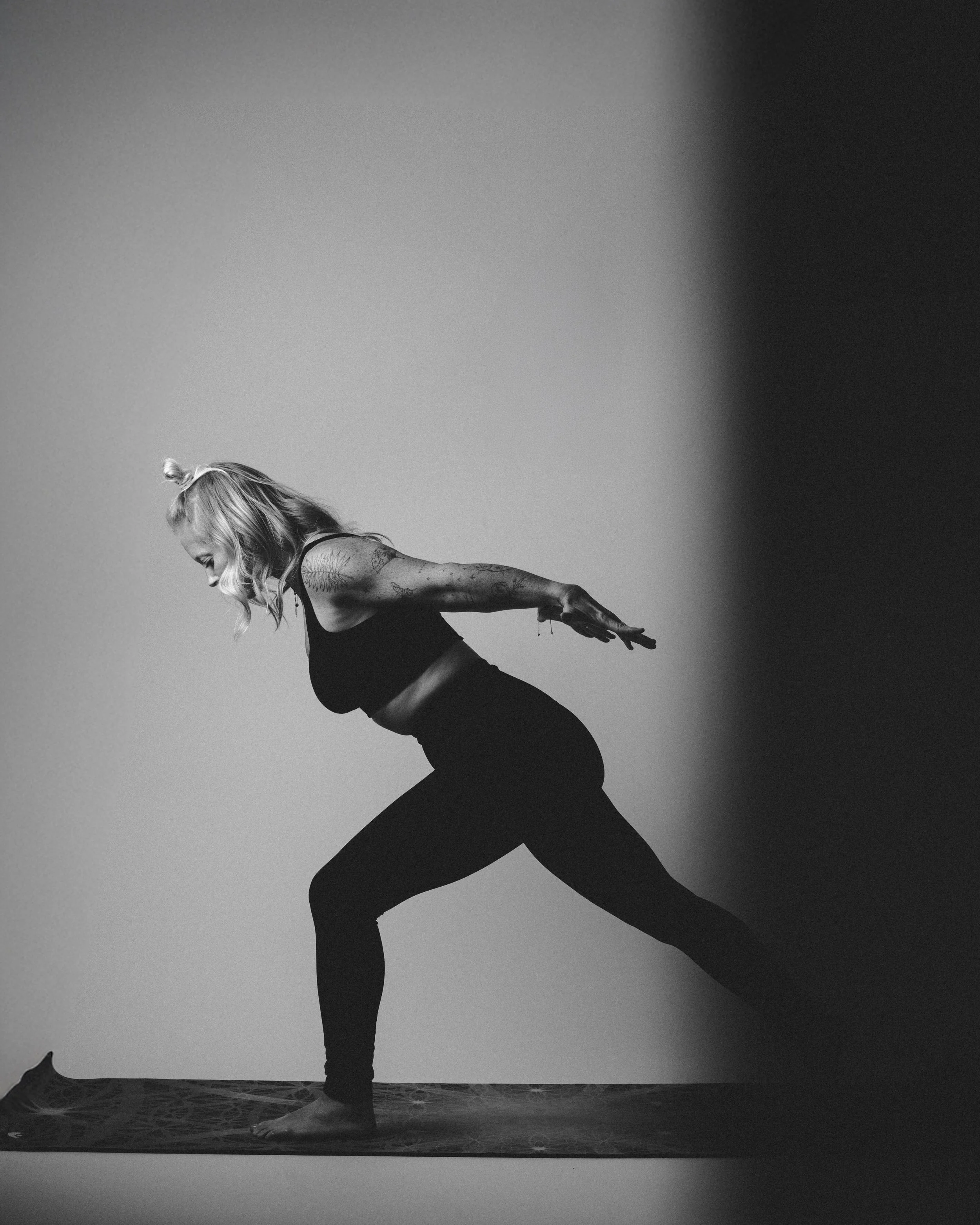 Black and white photo of a woman practicing yoga on a mat, in a Warrior pose, with a plain background.