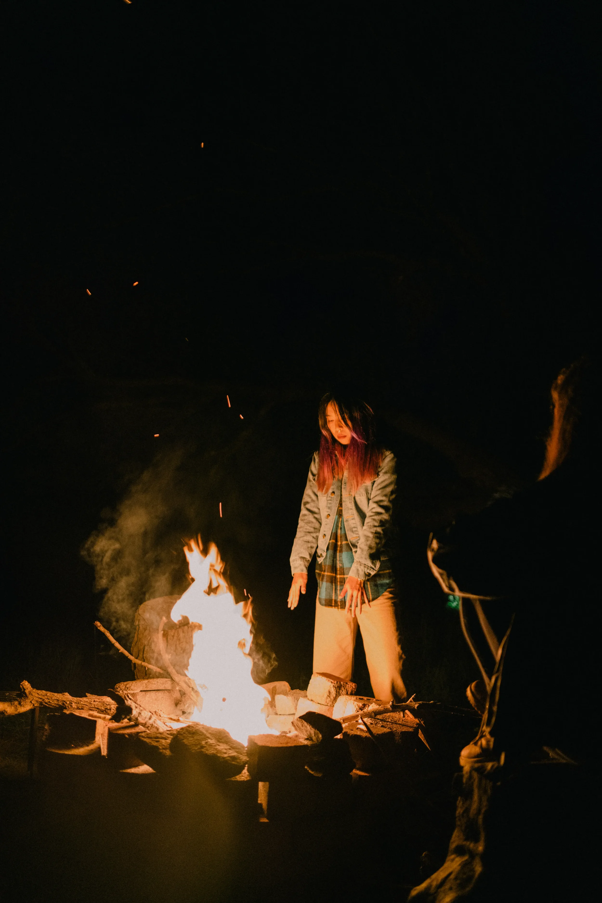 A woman stands near a campfire at night, surrounded by darkness.