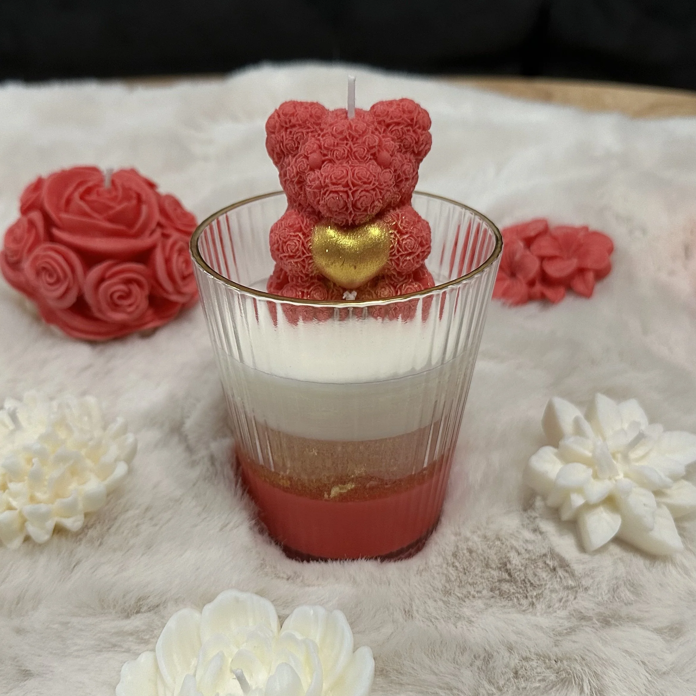 A teddy bear-shaped candle decorated with pink roses and a gold heart, placed inside a glass, surrounded by pink and white flower decorations on a soft, furry surface.