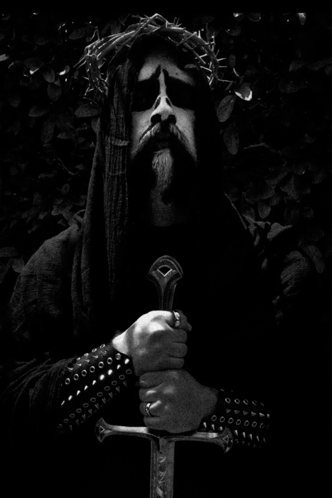 A person dressed as Jesus Christ with a crown of thorns, holding a sword with both hands, in front of dark foliage