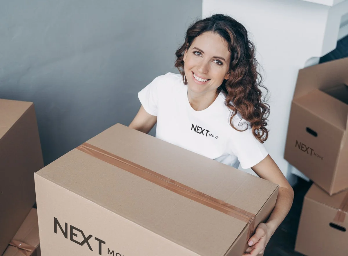 A smiling woman with curly brown hair holding a large cardboard box labeled 'Next Move' surrounded by other packed boxes.