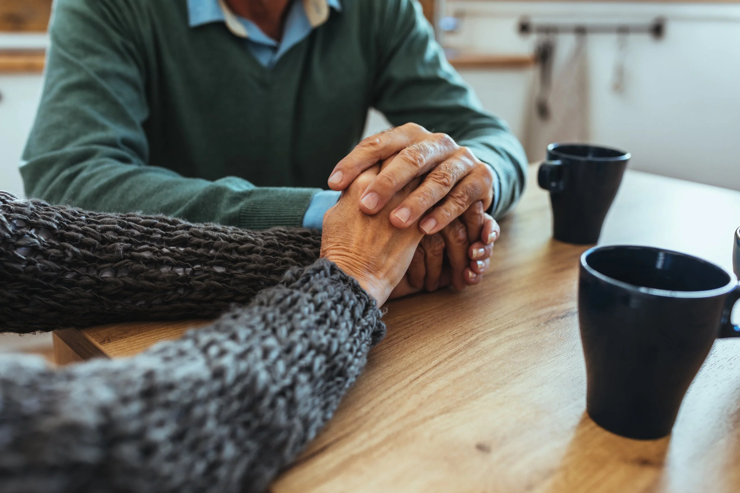 Two people holding hands across a wooden table with black mugs in the background.