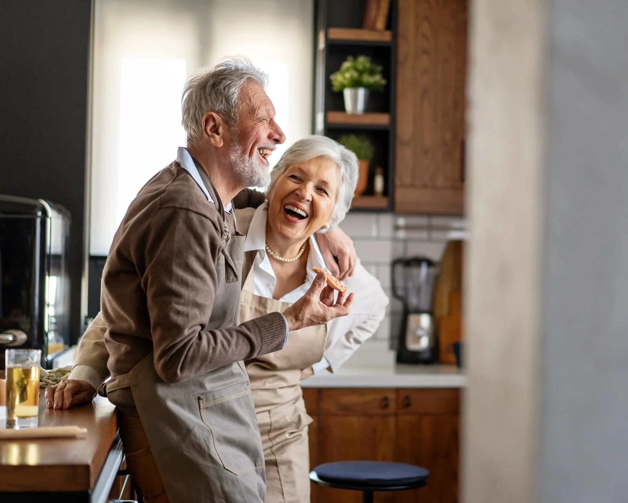 An elderly man and woman laughing and enjoying each other's company in a cozy kitchen, with the man holding a cookie.