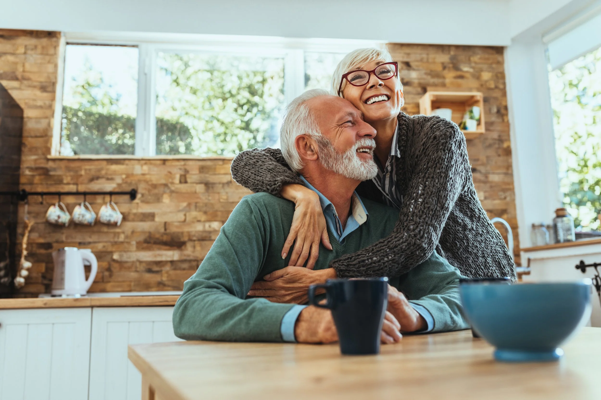 An elderly man with a white beard and wearing a green sweater is sitting at a kitchen table. A woman with short white hair, glasses, and wearing a gray sweater is hugging him from behind, both smiling joyfully. The background shows a modern kitchen with wooden accents and large windows letting in natural light.