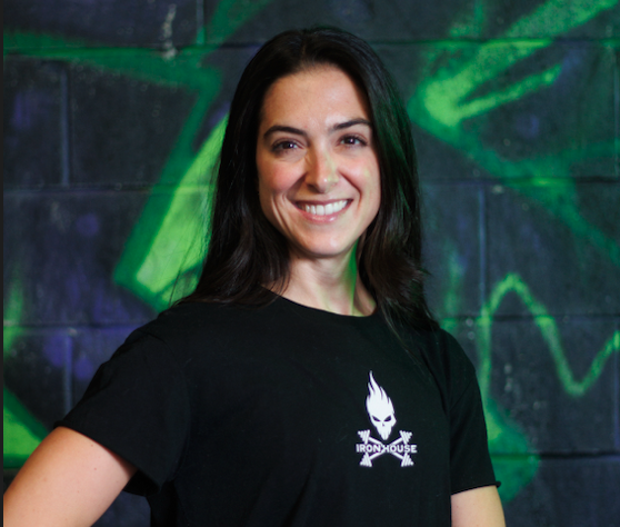 Young woman with dark hair smiling, wearing a black Ironhouse t-shirt, standing in front of a green and purple graffiti wall.