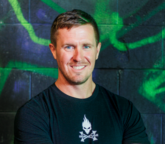 A smiling man with short brown hair wearing a black T-shirt with a white logo, standing in front of a graffiti-covered wall.