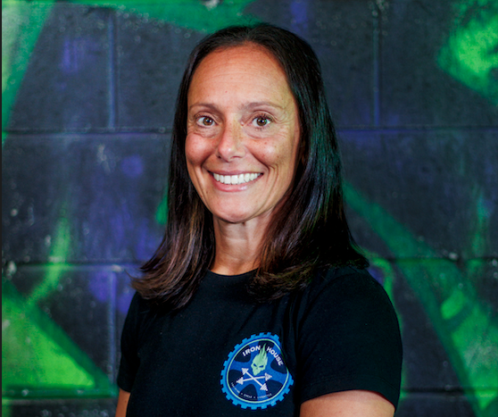 Woman with long dark hair smiling, wearing a black t-shirt with a blue and white logo, standing in front of a dark background with green and blue abstract lighting effects.