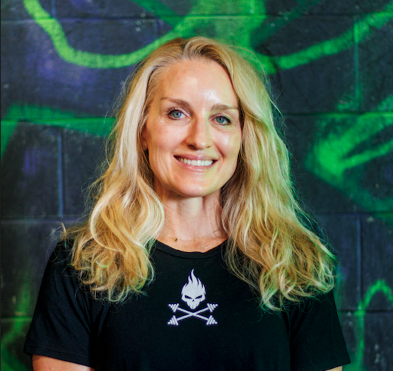 A woman with long blonde hair smiling, wearing a black T-shirt with a white flames and crossed arrows logo, standing in front of a dark wall with green graffiti.