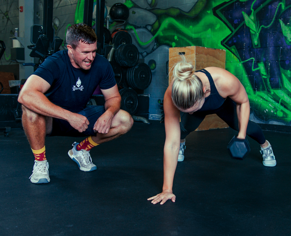 A woman in workout clothes doing a plank with a dumbbell, and a man coaching her in a gym with colorful mural walls.