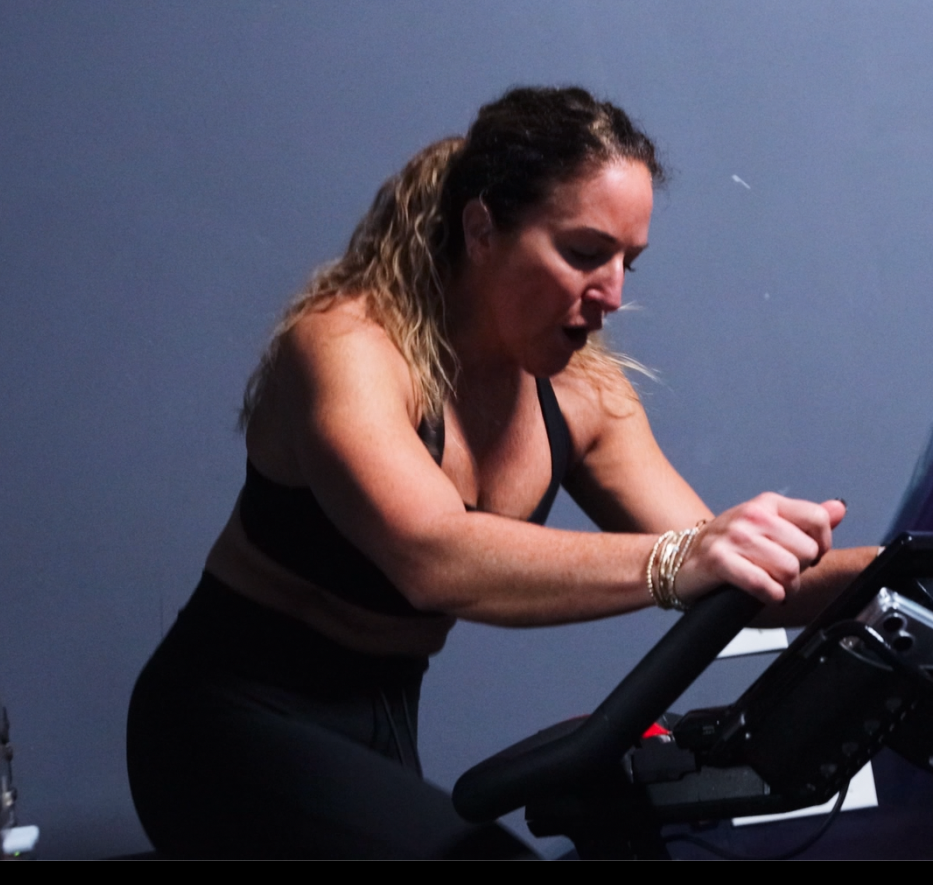 A woman with curly blonde hair in a black sports bra and bracelets on her wrist is exercising on a treadmill in a room with a dark blue wall.