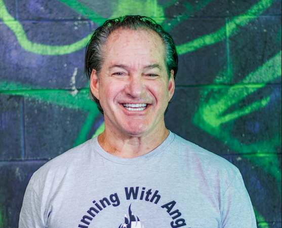 A smiling middle-aged man with slicked-back hair wearing a light gray T-shirt standing in front of a colorful graffiti background.