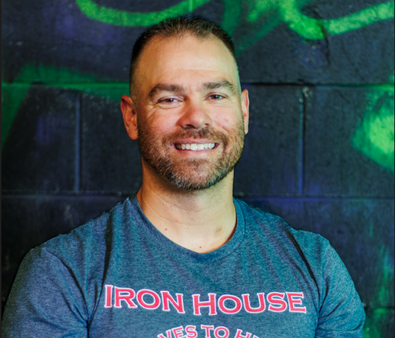 A smiling man with a beard and short hair, wearing a blue T-shirt that says 'Iron House' in front of a dark, graffiti-style background.