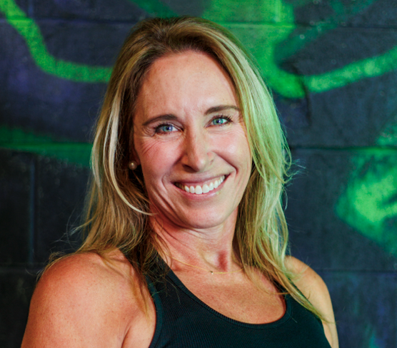 Smiling woman with blonde hair wearing a black tank top, standing in front of a dark wall with green graffiti.