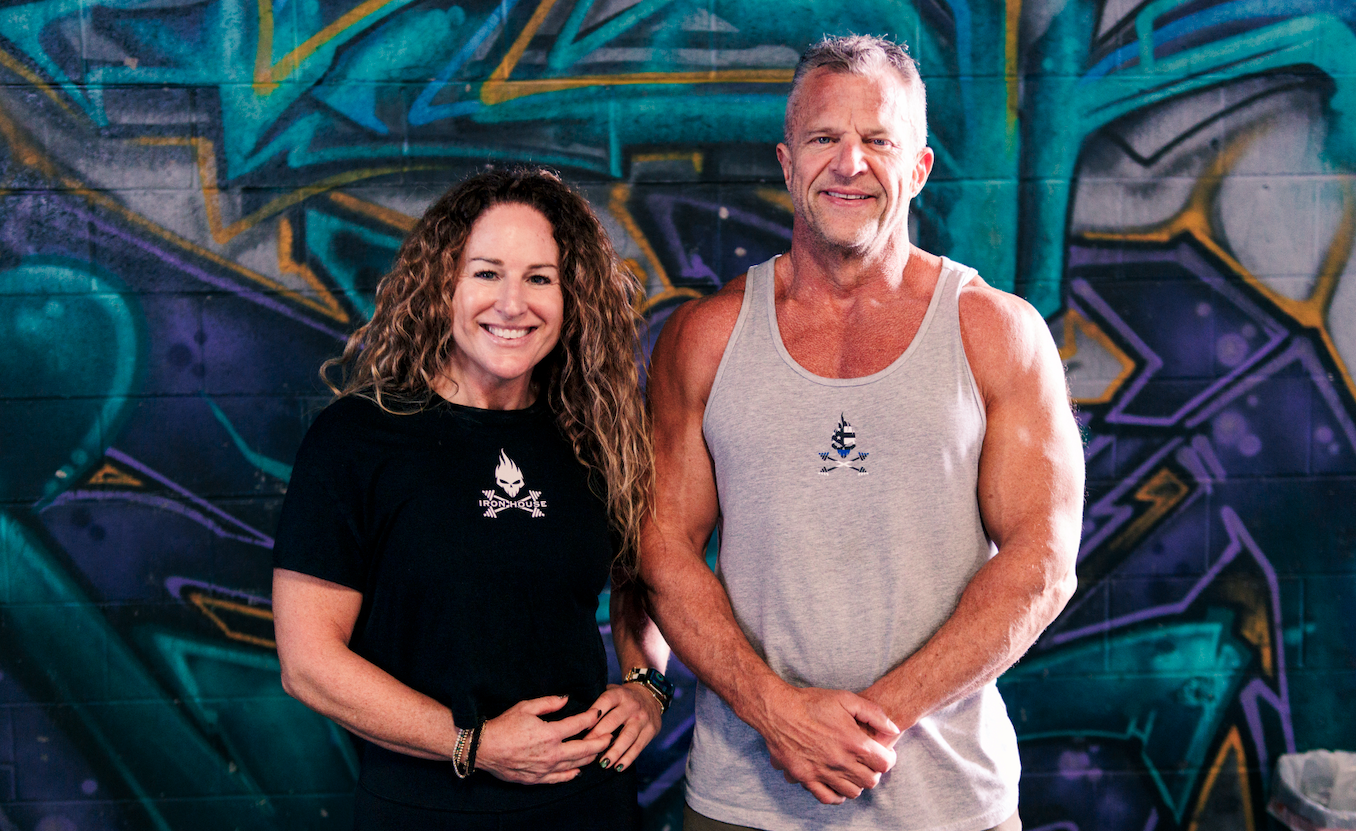 A smiling woman and man stand in front of a colorful graffiti wall. The woman has curly hair and wears a black t-shirt with a logo, while the man has short gray hair and wears a gray tank top with a small logo.