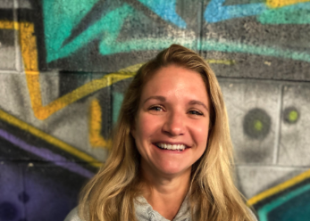 A smiling woman with blonde hair standing in front of graffiti-covered wall.