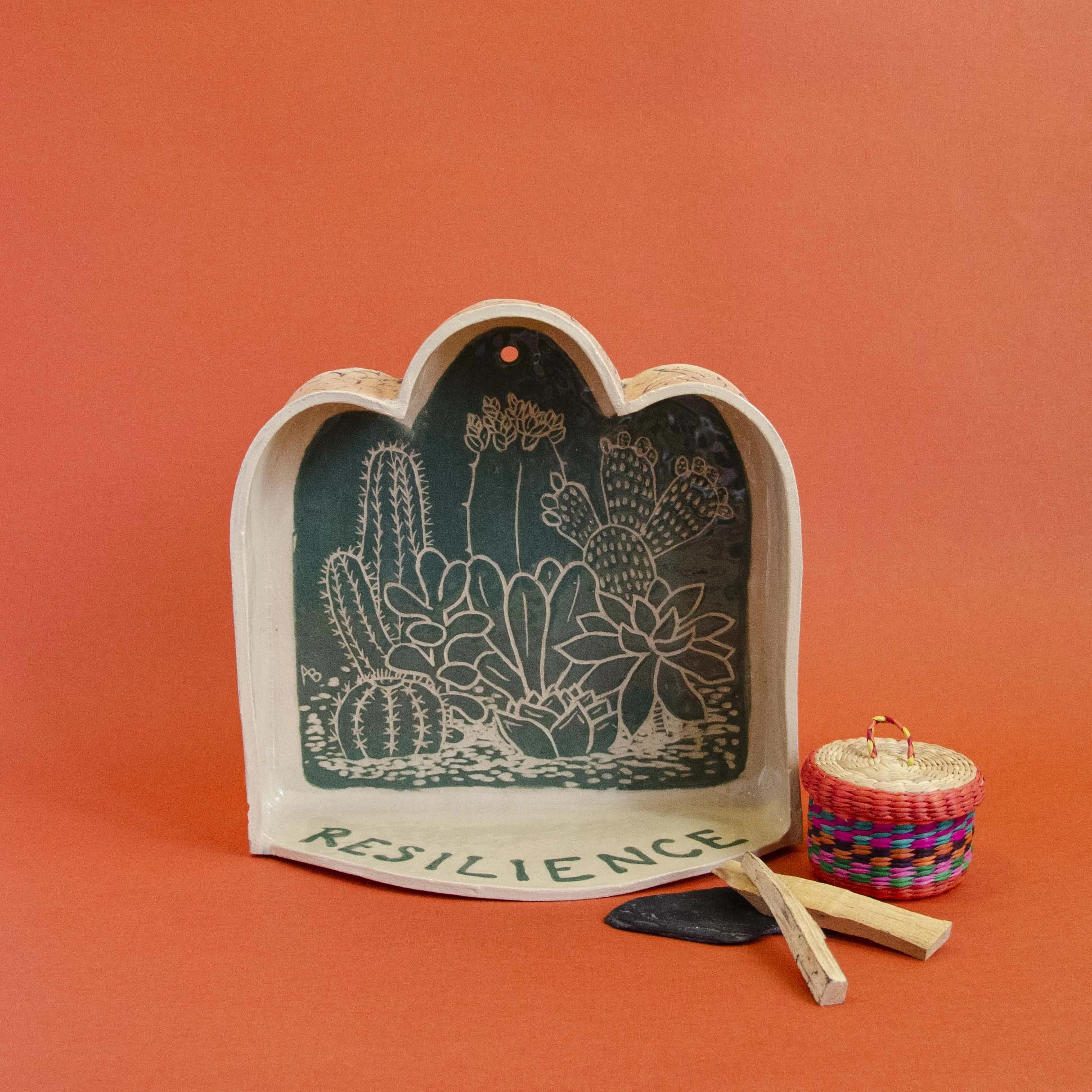 A decorative black and white reverse-painted ceramic dish with a desert cactus design and the word 'RESILIENCE' written on the bottom edge, placed against an orange background. To the right, a colorful woven basket with a lid, a small black stone, and two pieces of wood.