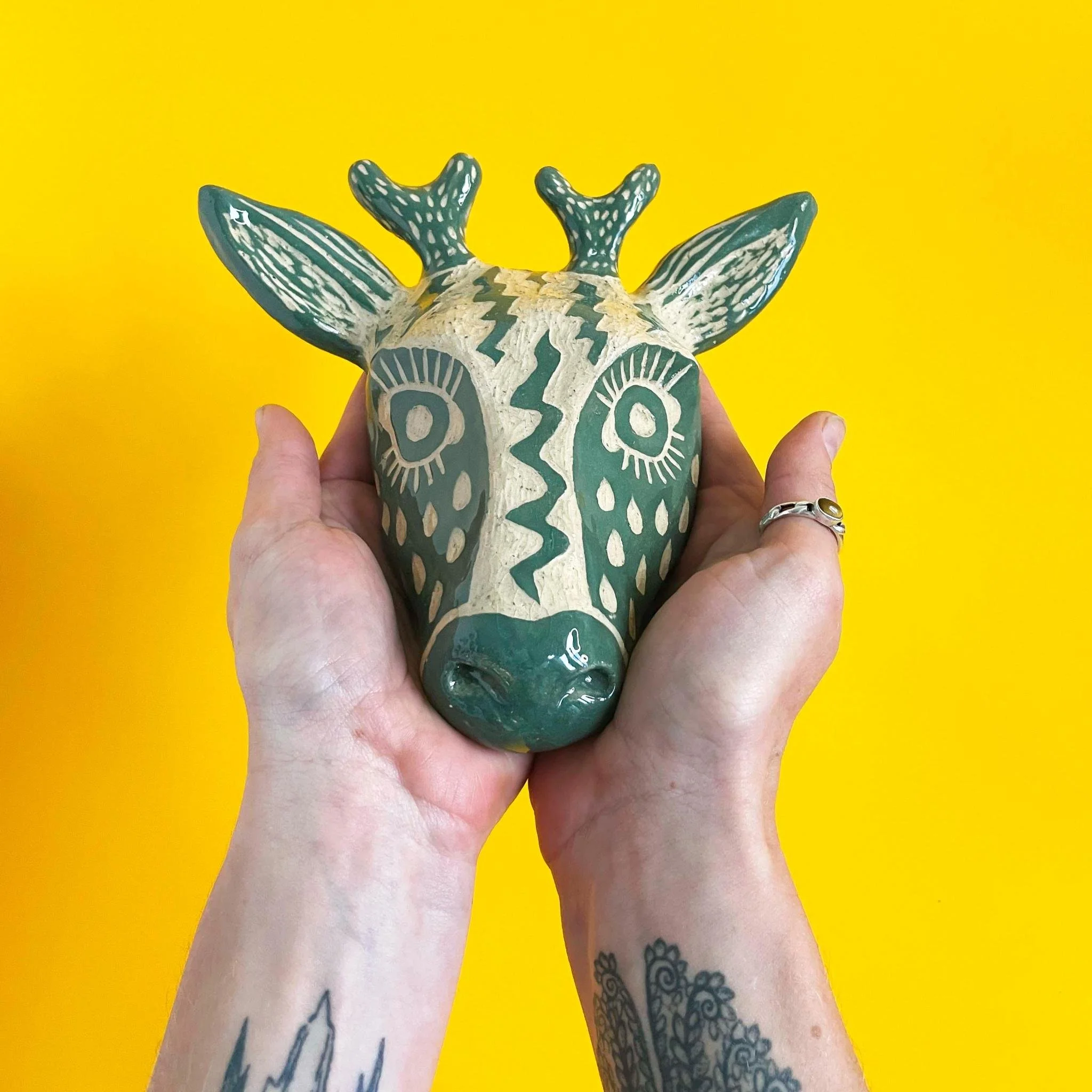 Person holding a decorated ceramic deer head sculpture with a yellow background.