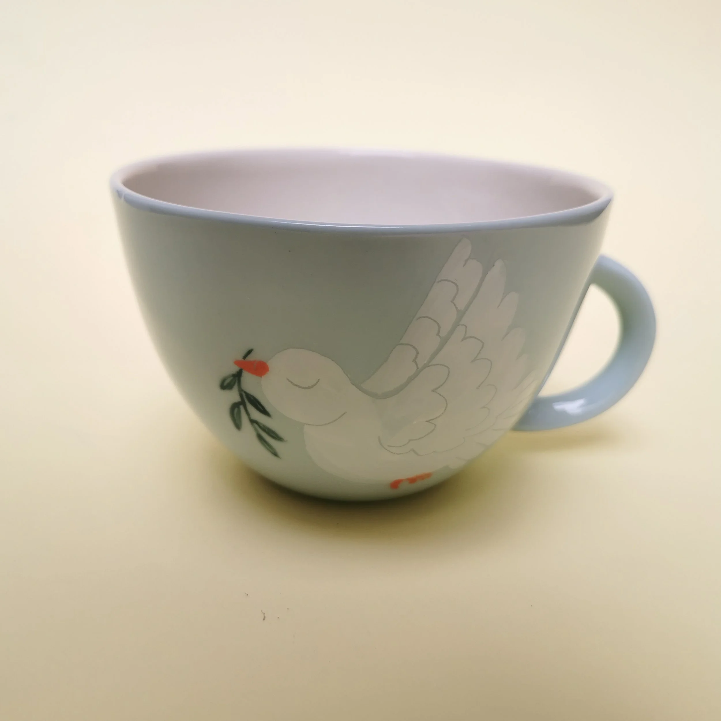 White ceramic cup with a dove design holding an olive branch painted on it, set against a pale yellow background.