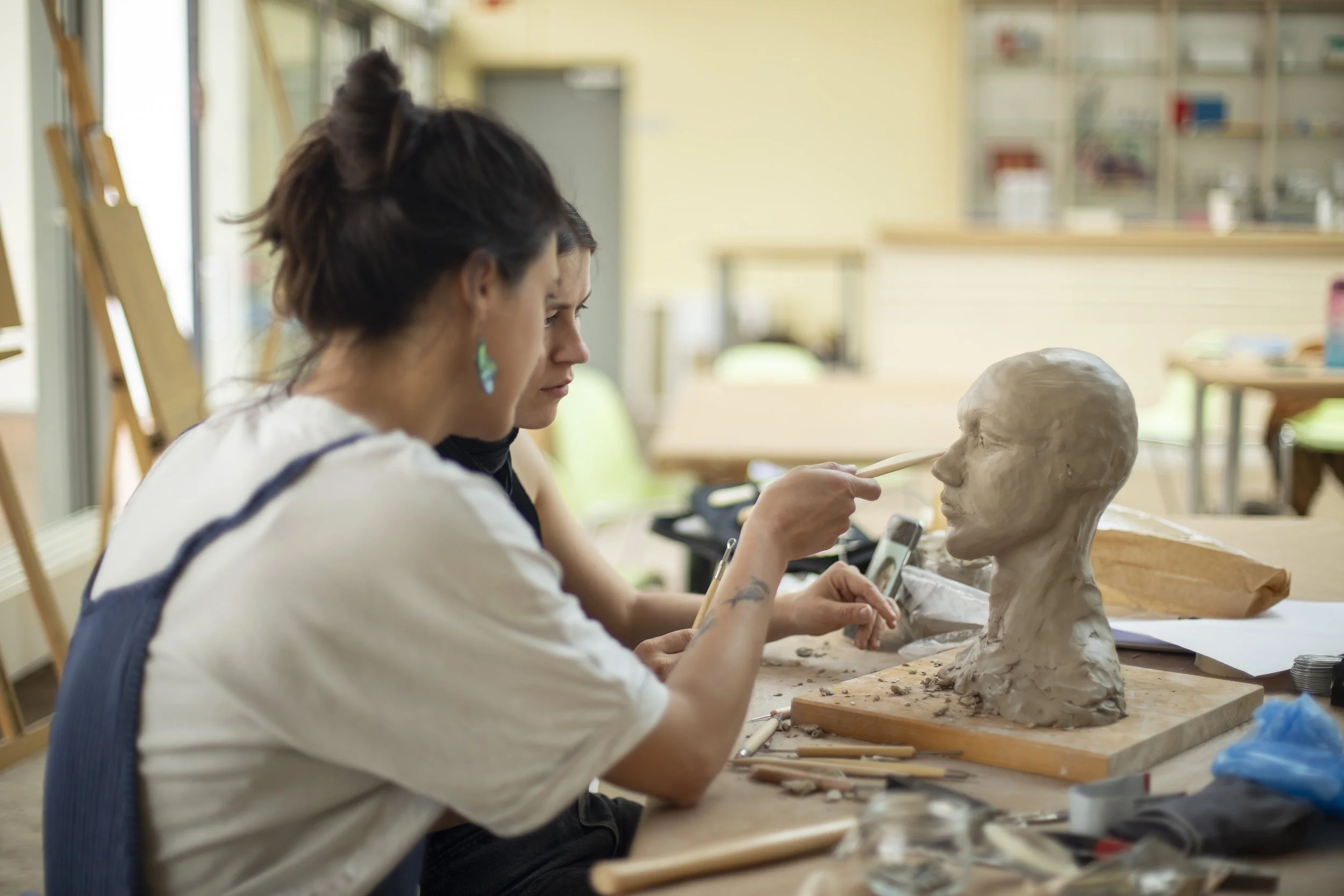Two women sculpting a clay bust of a person's head in an art studio.
