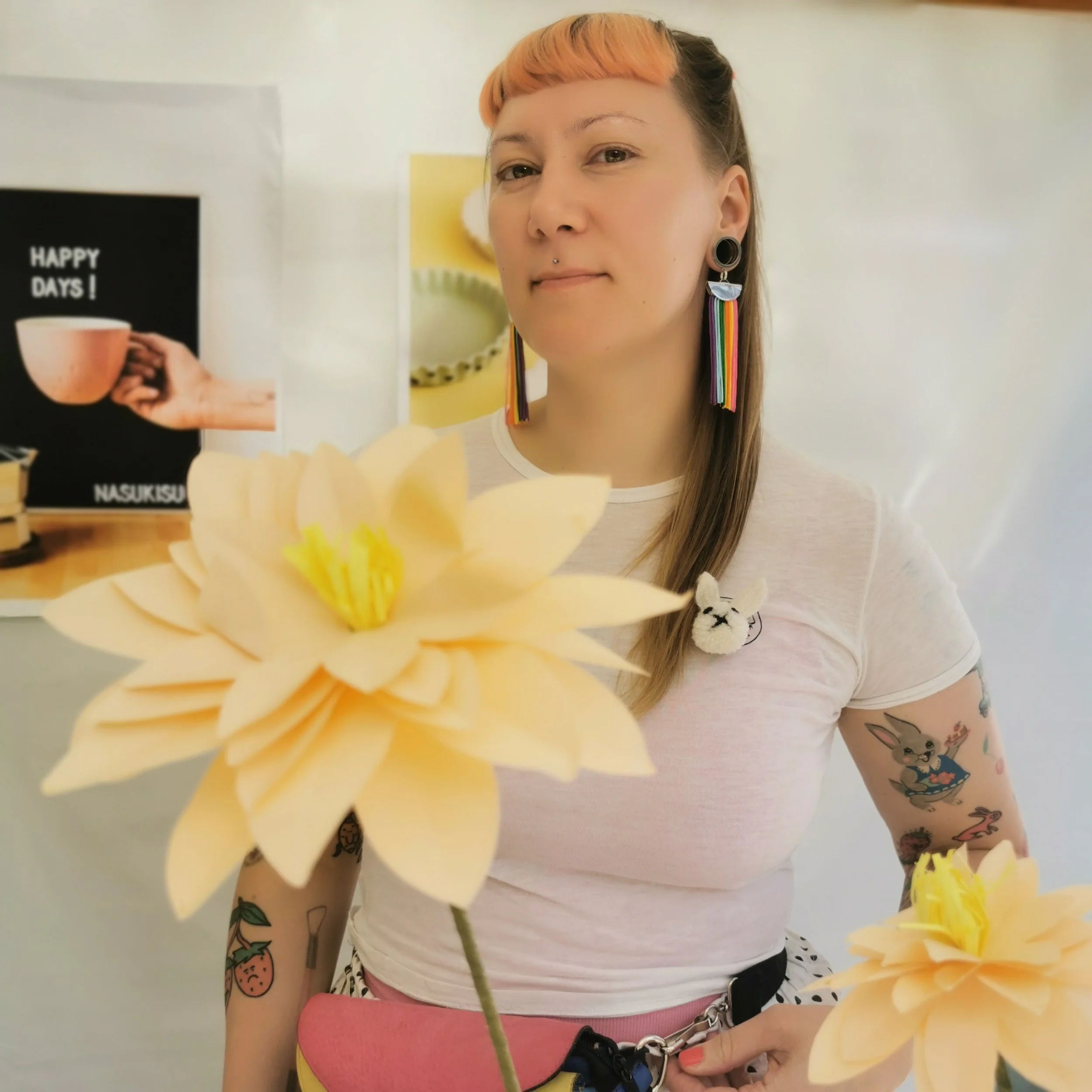 A woman with colorful earrings and tattoos, holding a yellow flower in front of her, in a casual indoor setting.