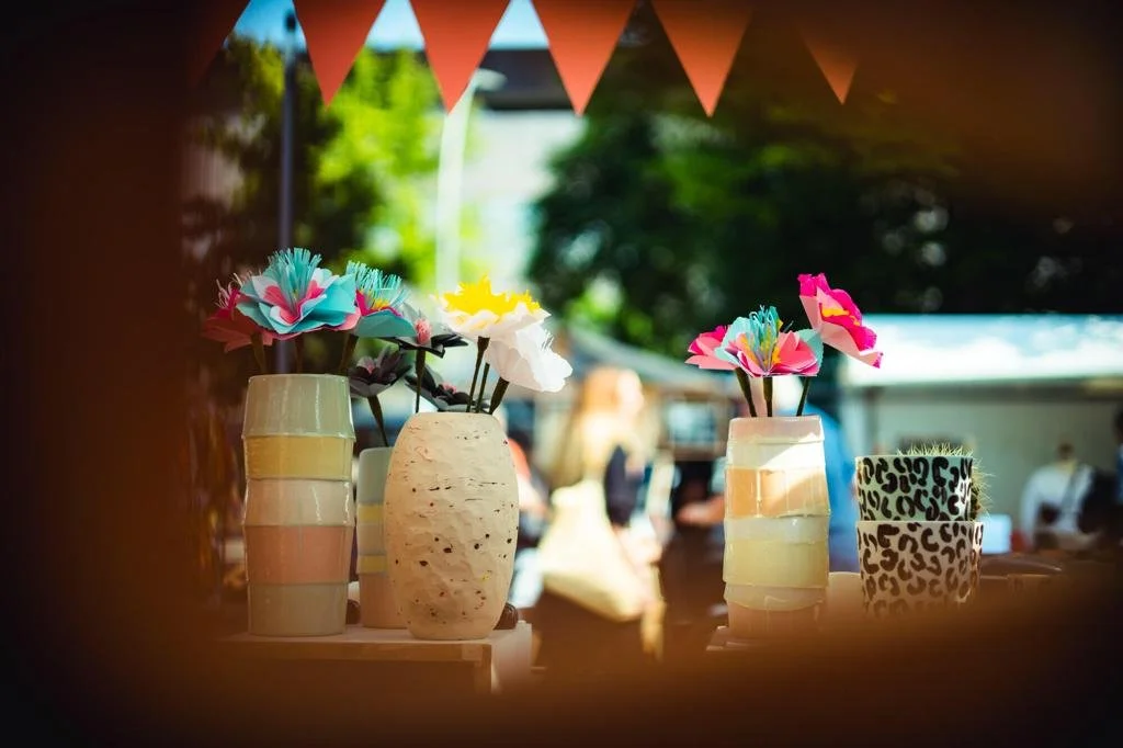 Colorful paper flower decorations in various vases and cups at an outdoor event with trees and people in the background.