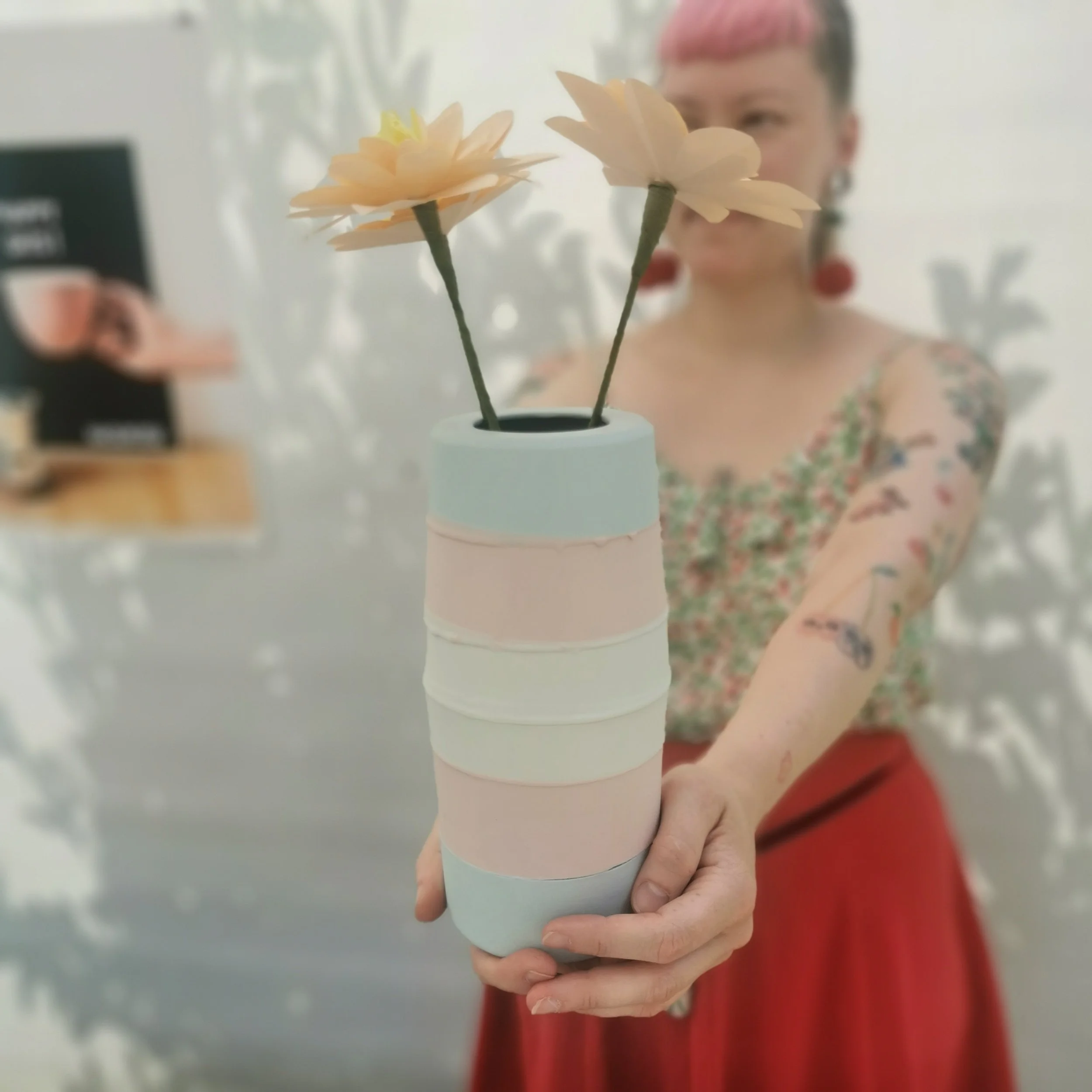 A woman with pink hair and tattoos holding a stack of pastel-colored cups with two pink flowers in a vase on top.