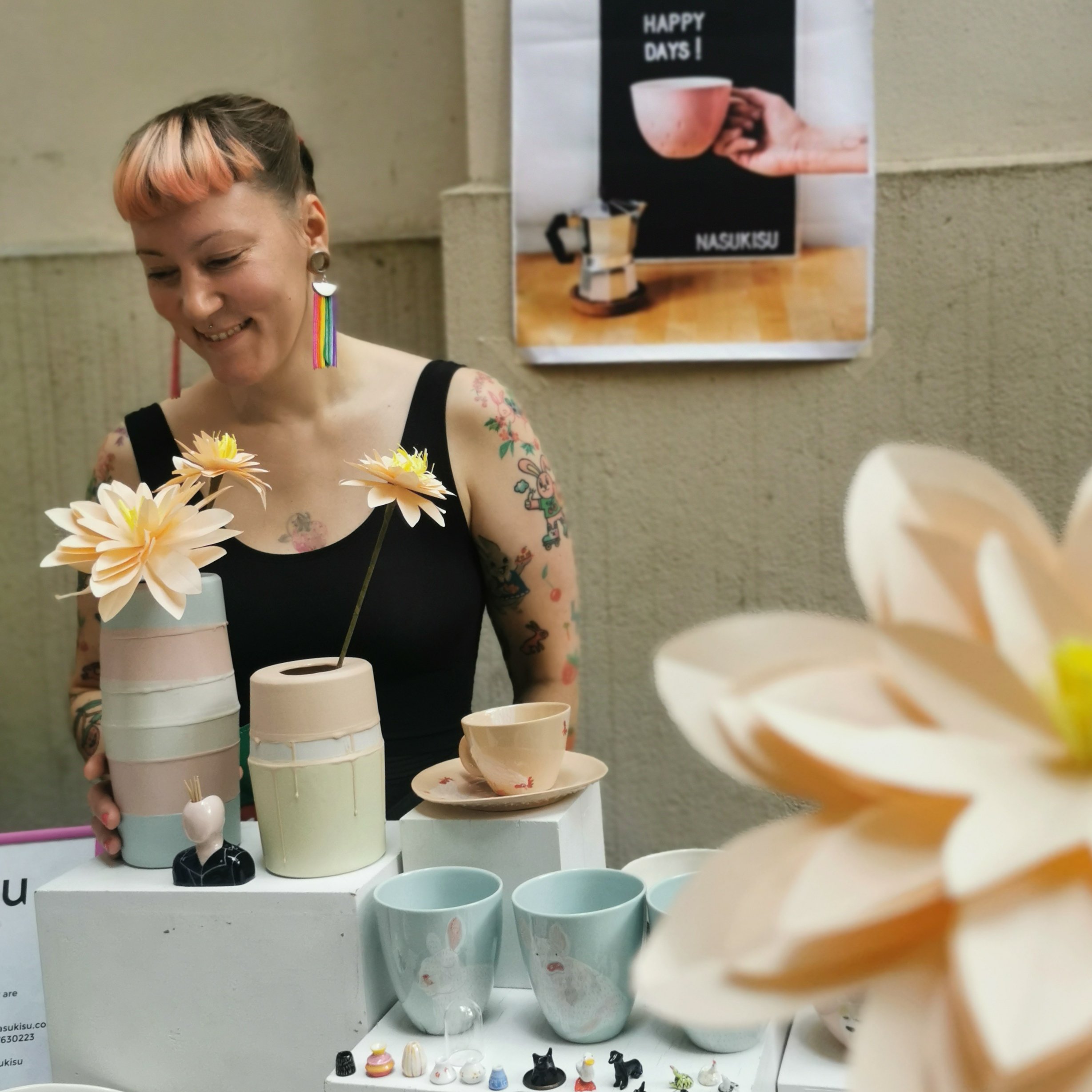 A woman with pink and blonde hair, tattoos, and rainbow earrings stands behind a display of pastel-colored ceramic vases, cups, and decorative items with artificial flowers in the foreground. She is smiling and looking downward.