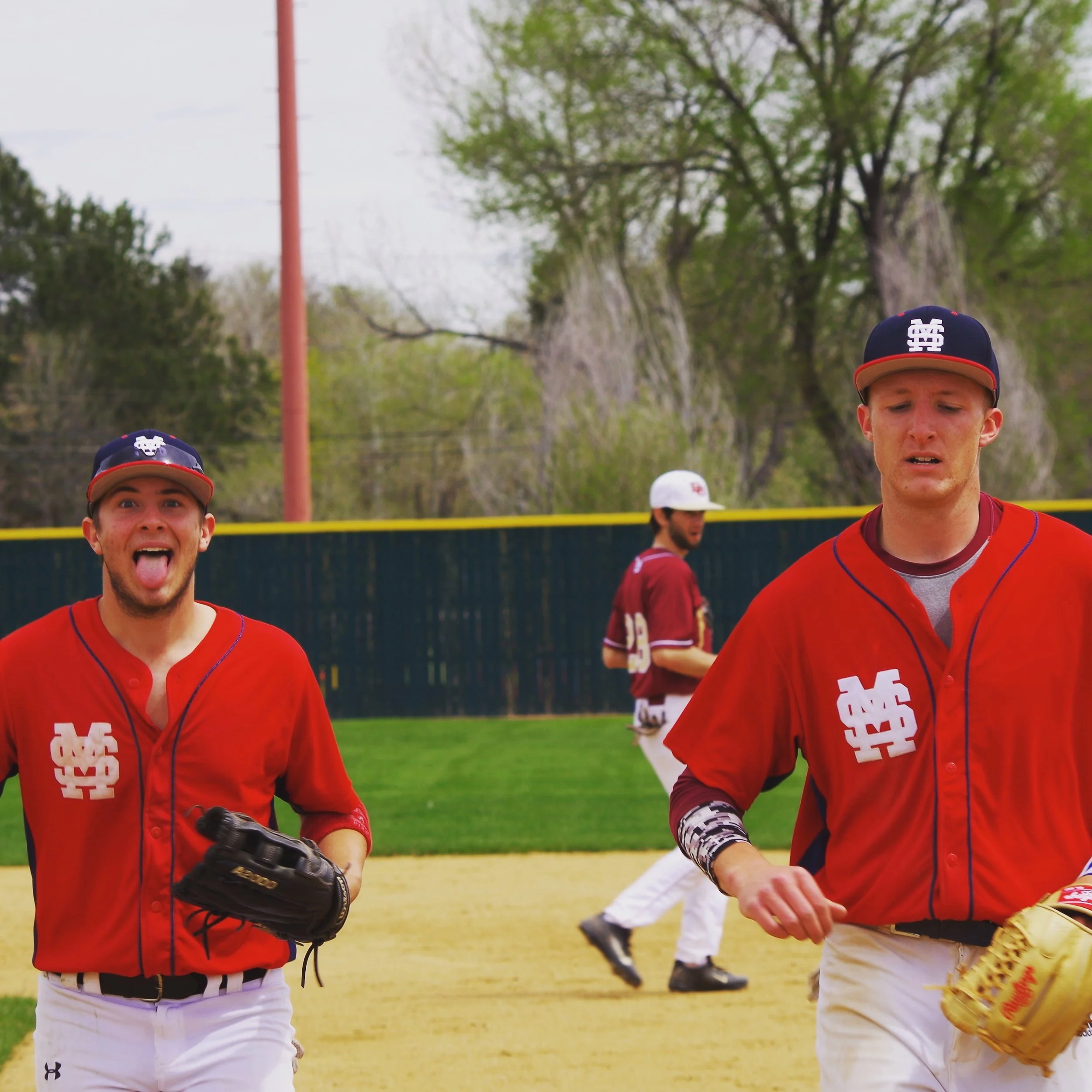 Two baseball players in red jerseys with black caps on a baseball field, one making a silly face with tongue out and the other looking serious, with another player in the background walking.