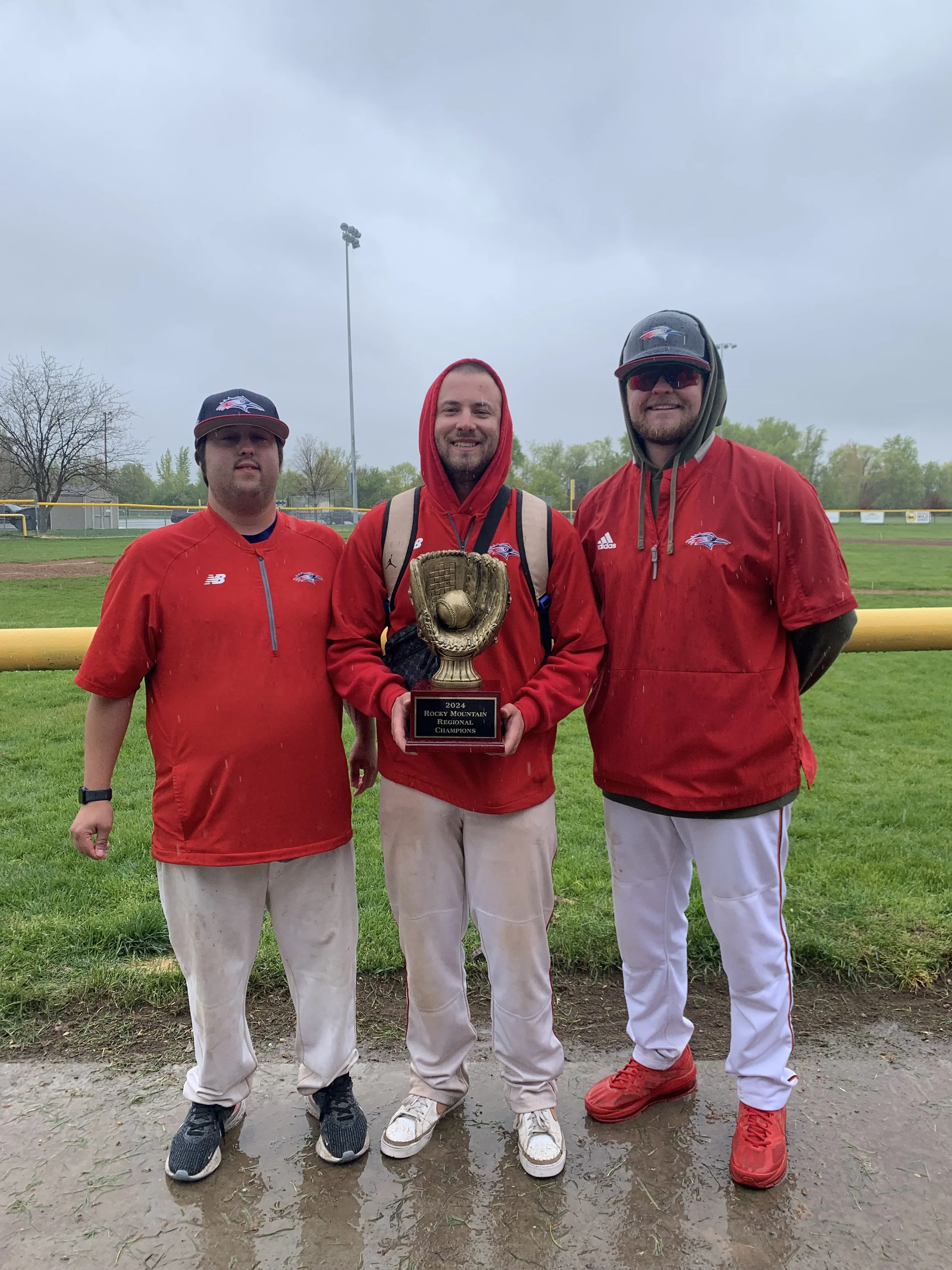 Three men in baseball gear standing on a field, holding a trophy.