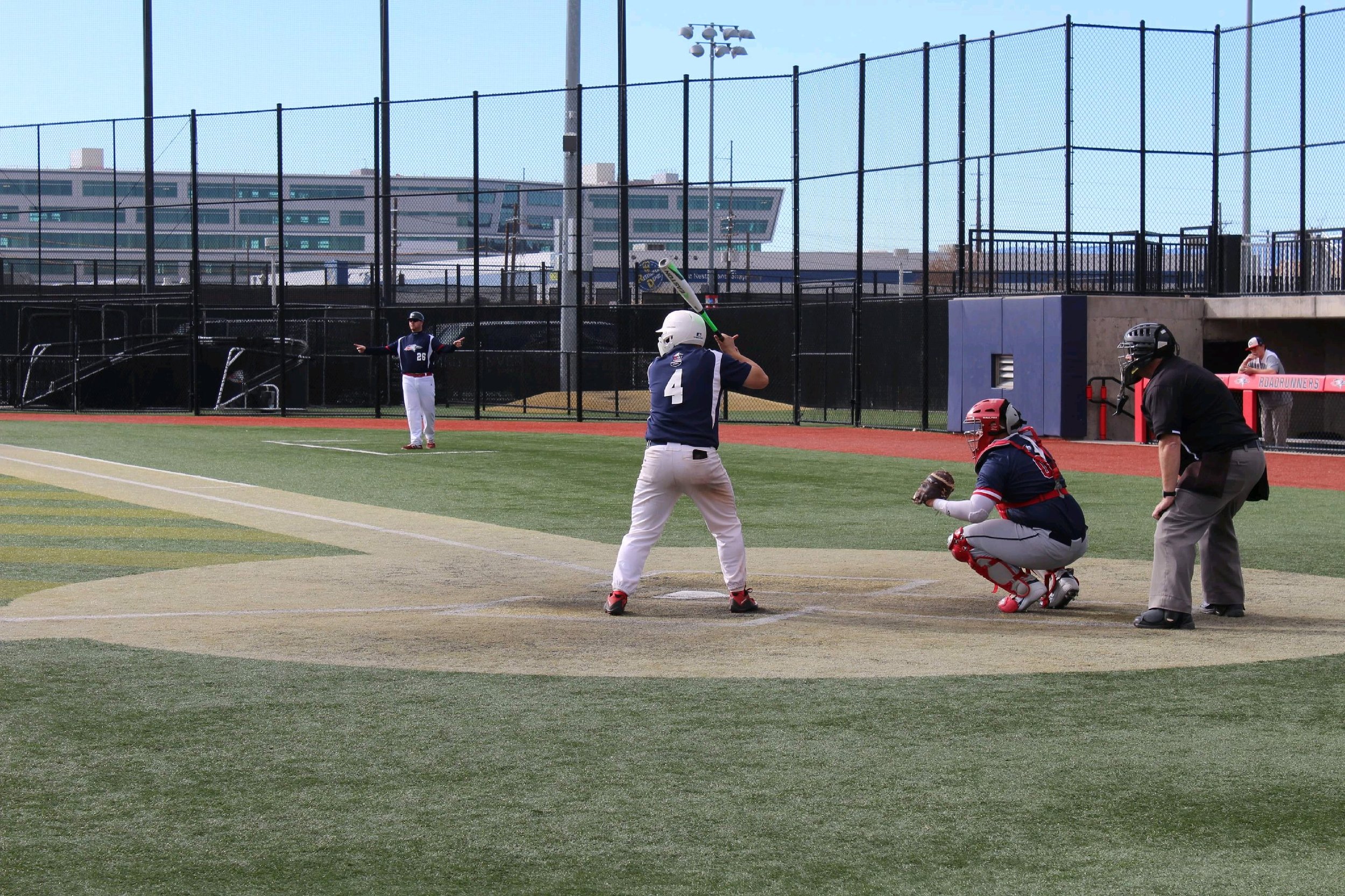 Young baseball players in a game, with a batter at the plate, catcher and umpire behind him, and a player standing in the field, on a baseball field with a fence and buildings in the background.