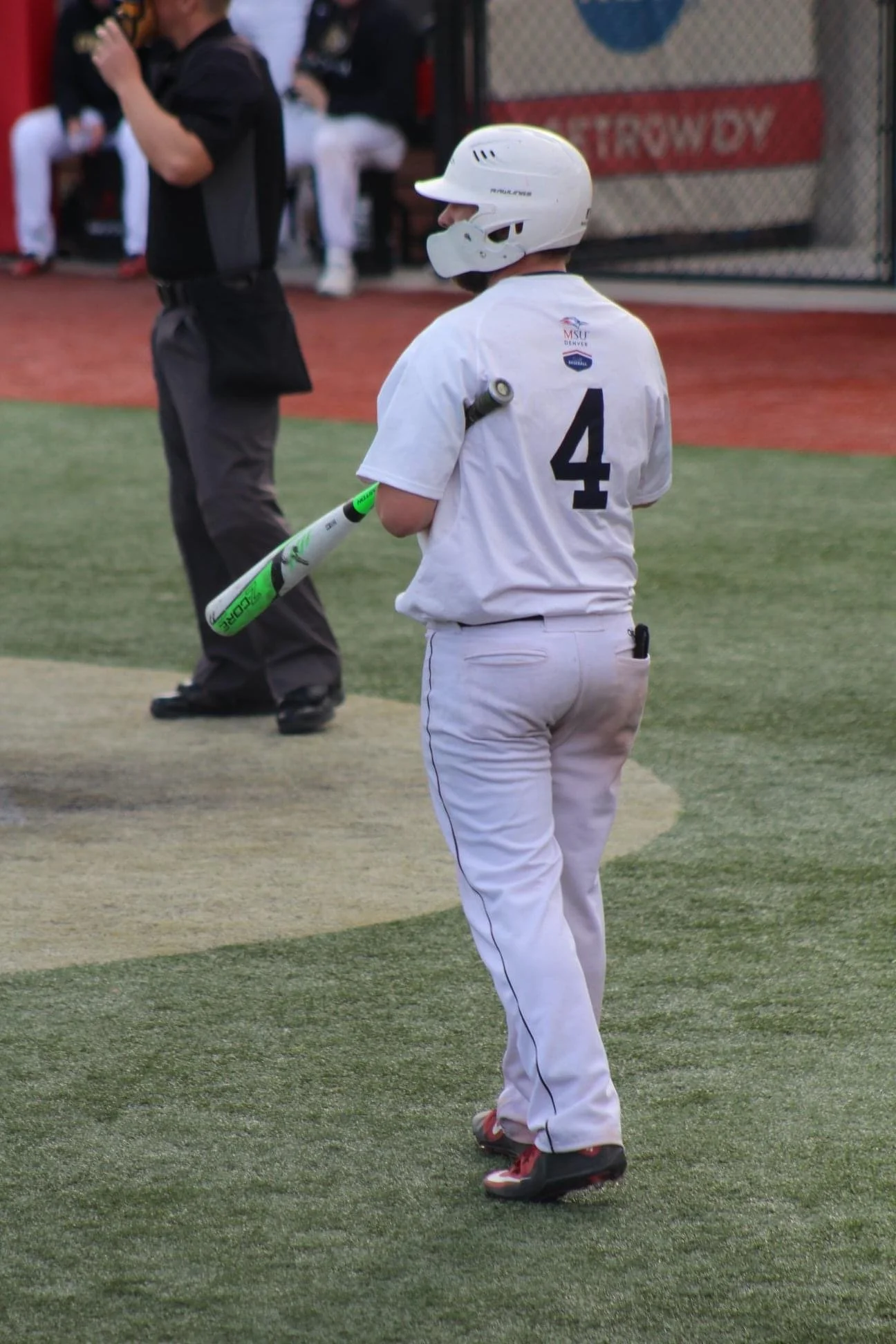 A baseball player in a white uniform with the number 4 and MSU logo on the back, holding a green and white bat, standing on the field with an umpire and other players in the background.
