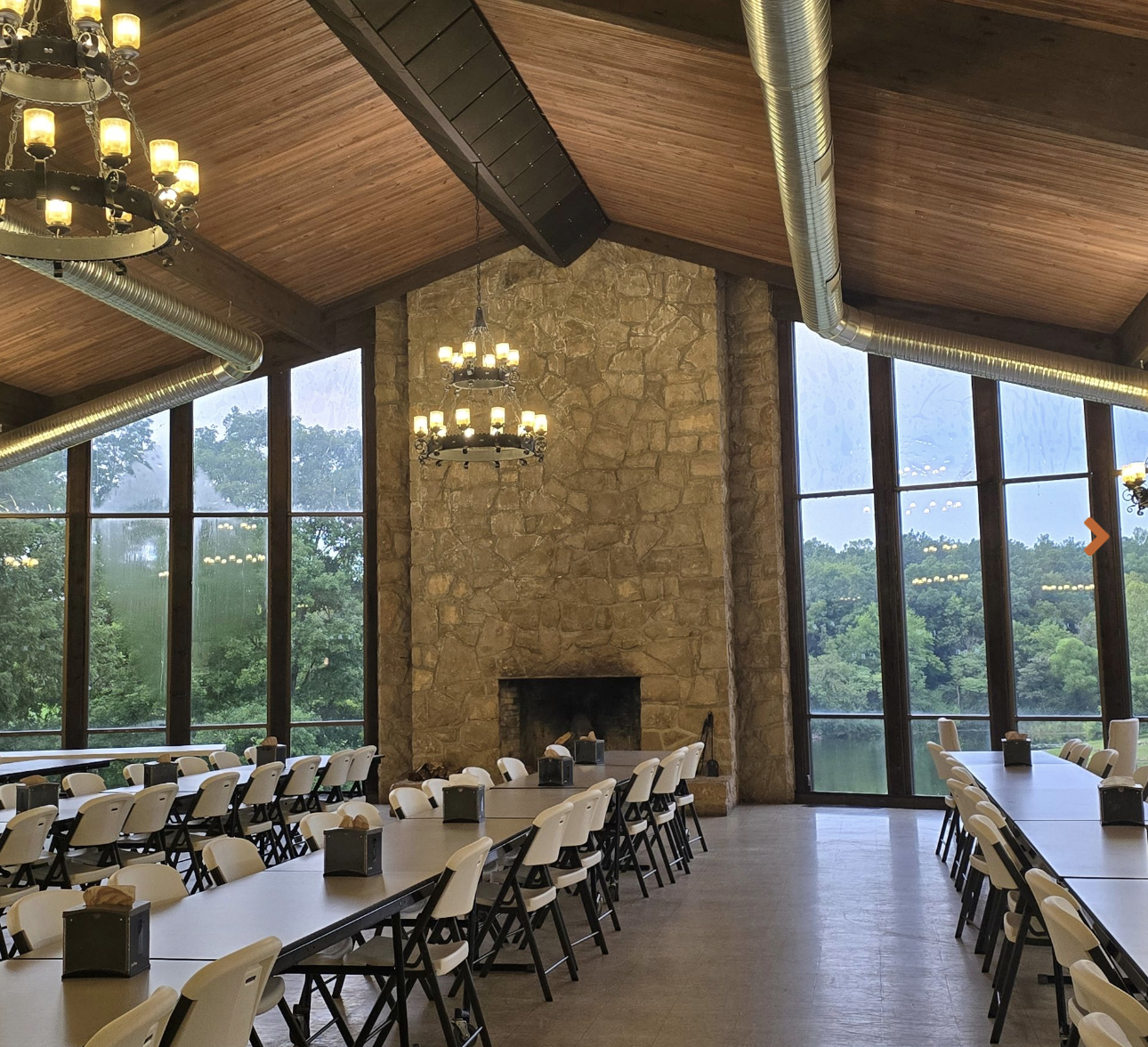 Interior of a spacious dining area with long tables and white chairs, large windows overlooking green trees, a stone fireplace, and chandeliers hanging from a wooden ceiling.